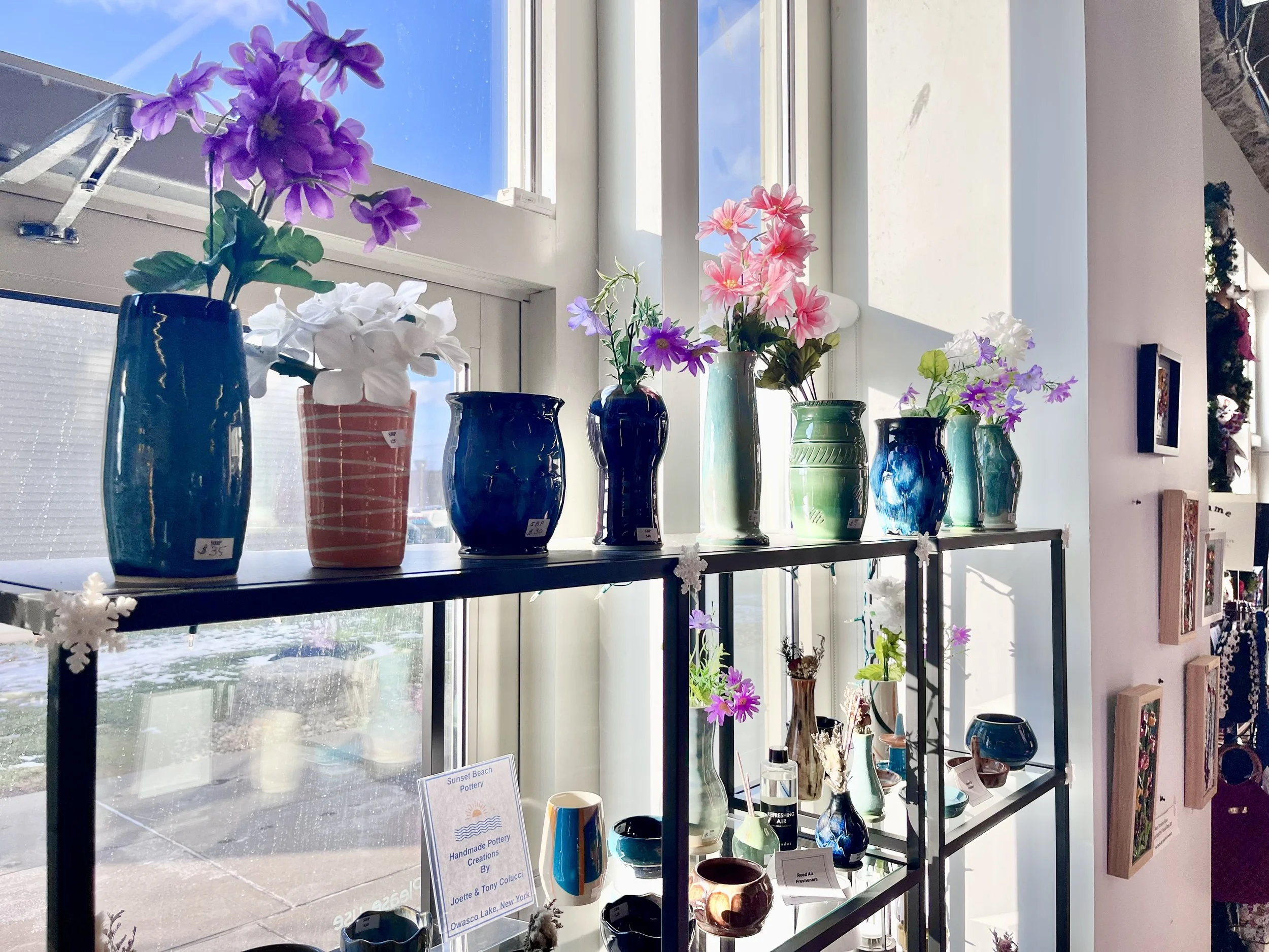 Sunset Beach pottery display with colorful vases and artificial flowers near window, with sunlight illuminating the scene.