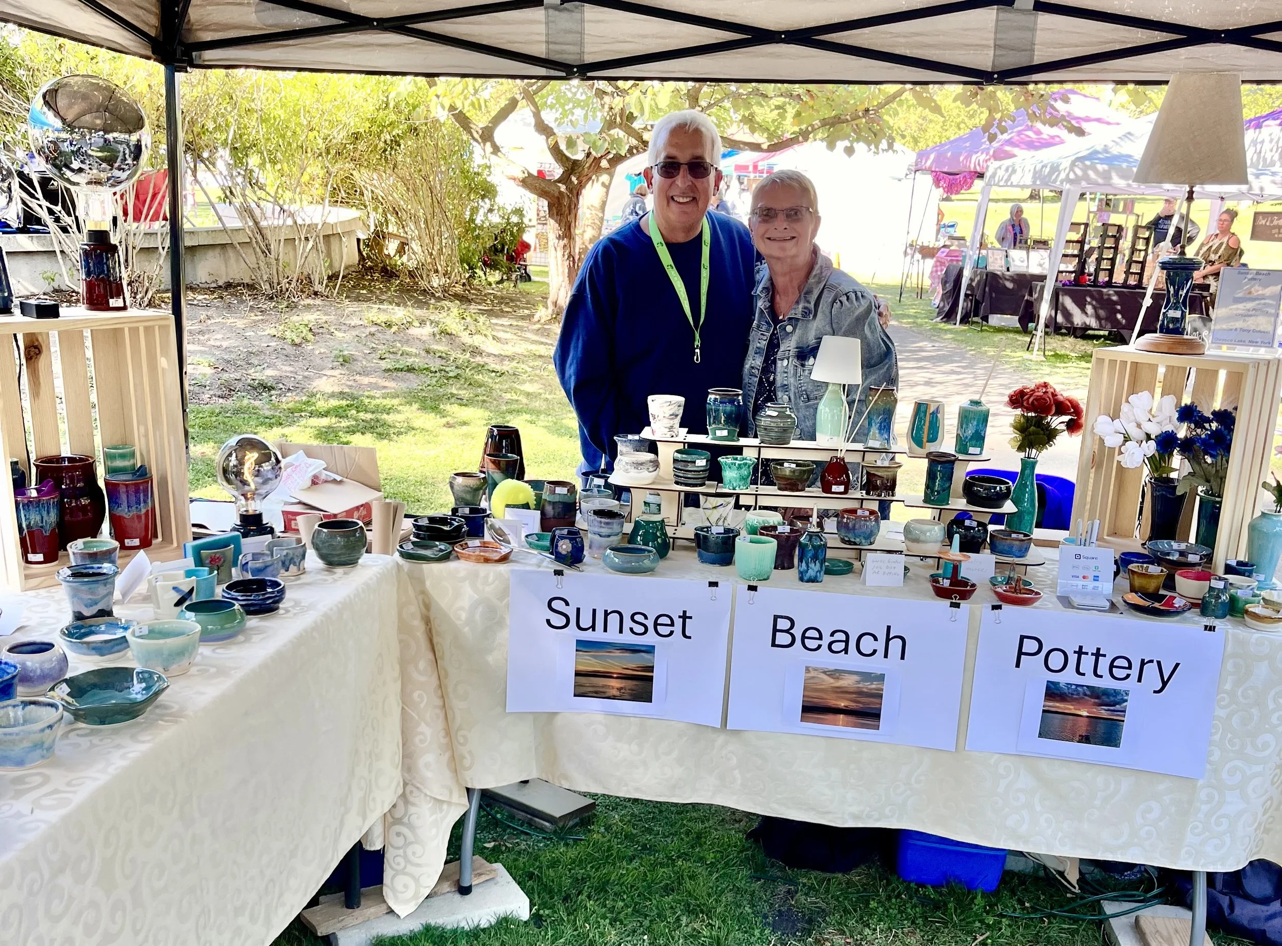 A woman and a man standing behind a table displaying colorful pottery at an outdoor market. The table is decorated with signs that read "Sunset," "Beach," and "Pottery" with sunset images underneath. Other vendor booths and tents are visible in the background.