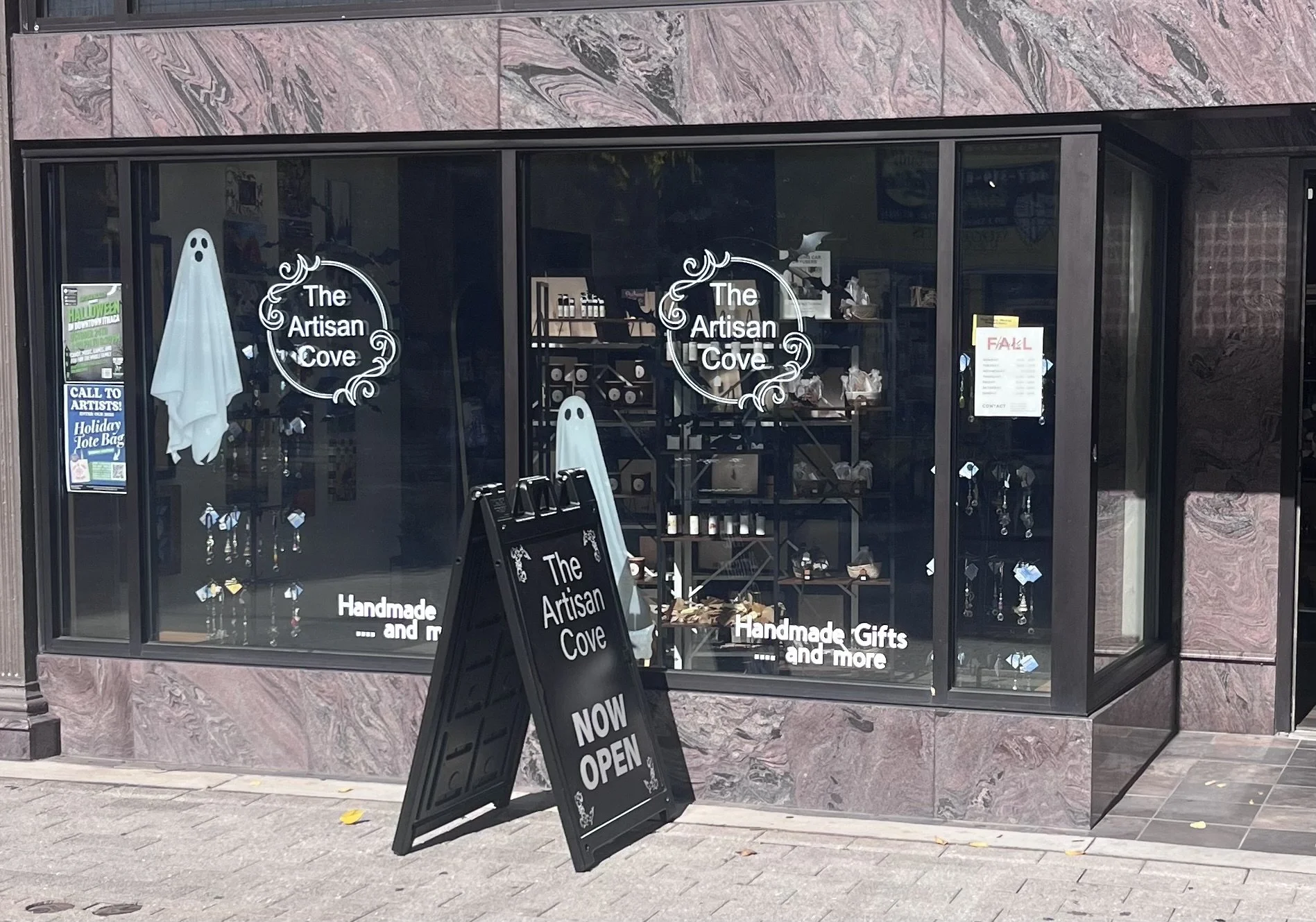 Storefront window for The Artisan Cove, featuring signs that say "Now Open" and "Handmade Gifts and More." The window displays various handmade items and jewelry, with a Ghost decoration and a sidewalk sign outside.
