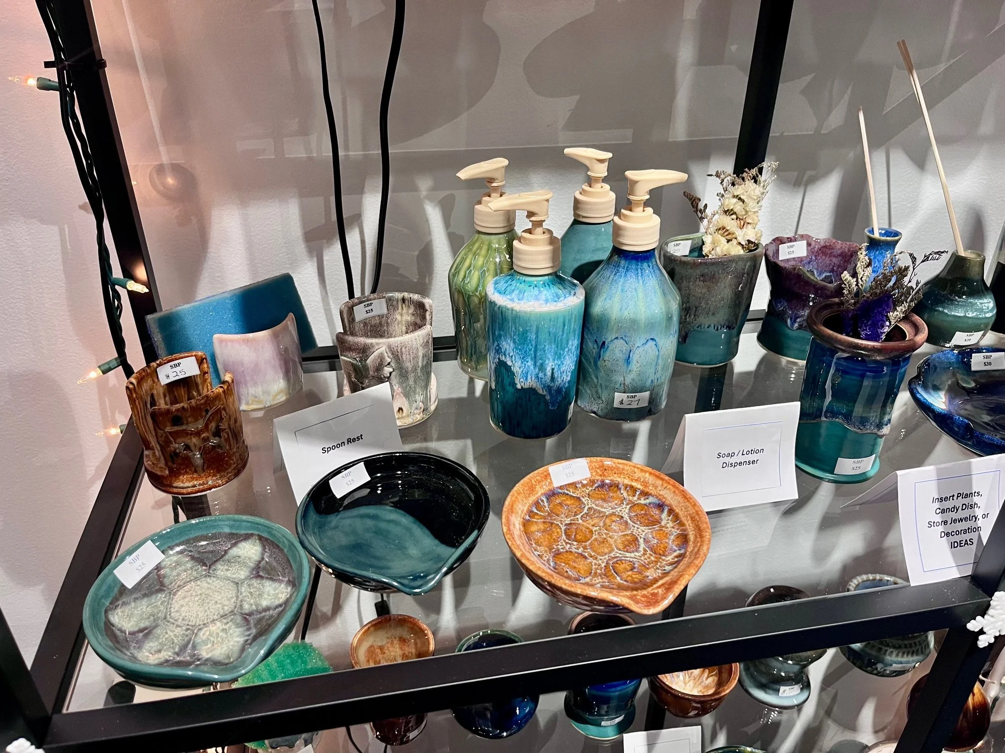 A display of various ceramic items on a glass table including soap dispensers, cups, bowls, and decorated pieces, with price tags and labels for each item.