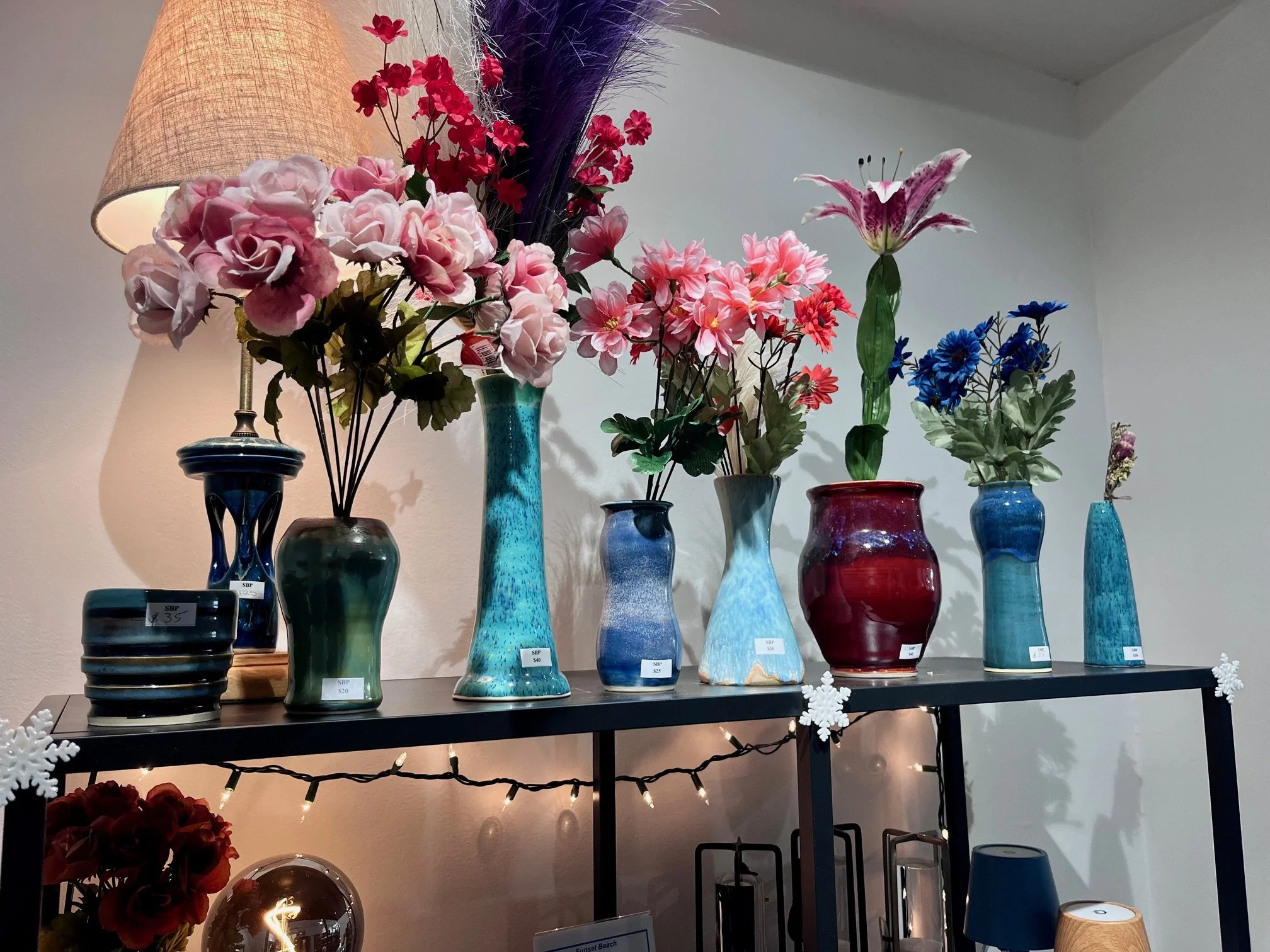 Display of colorful ceramic flower vases with artificial flowers, arranged on a black shelf with a lamp and string lights in the background.