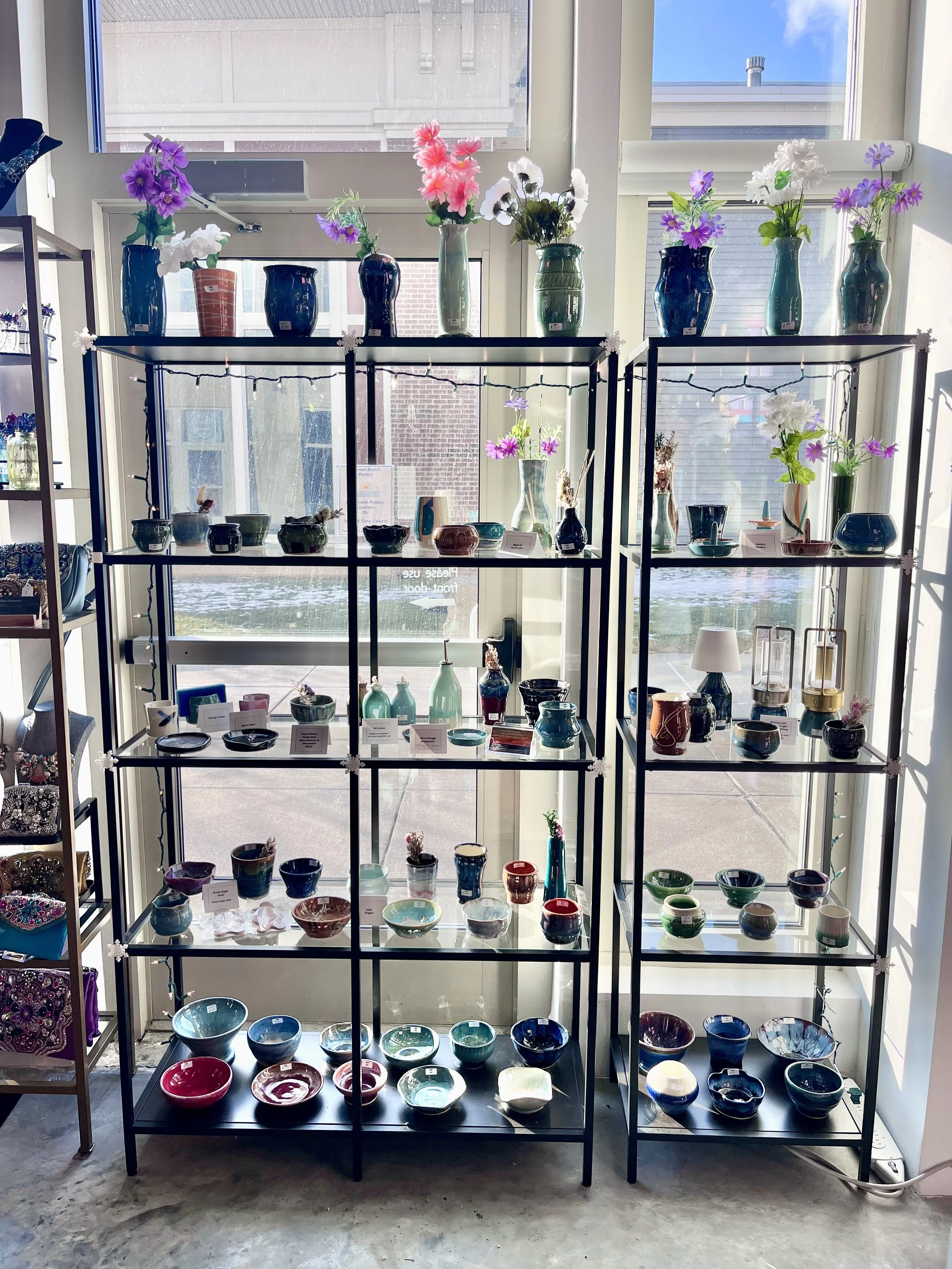 Display of various ceramic bowls and vases on black metal shelves with LED string lights, nearby a window with flowering vases on top.