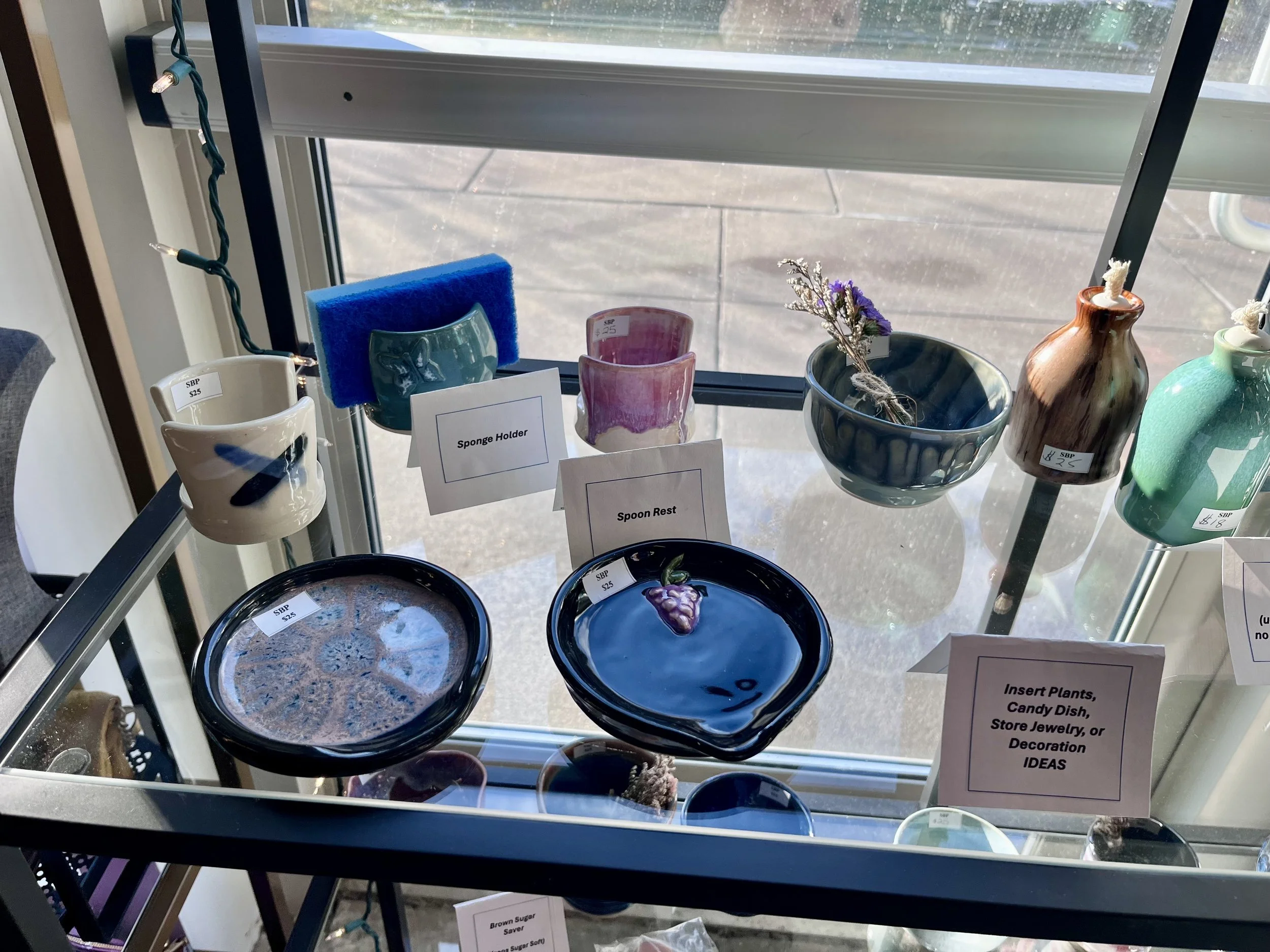 Display of decorative ceramic and glass bowls, mugs, and vases on a glass table near a window, with small signs indicating their uses and prices.