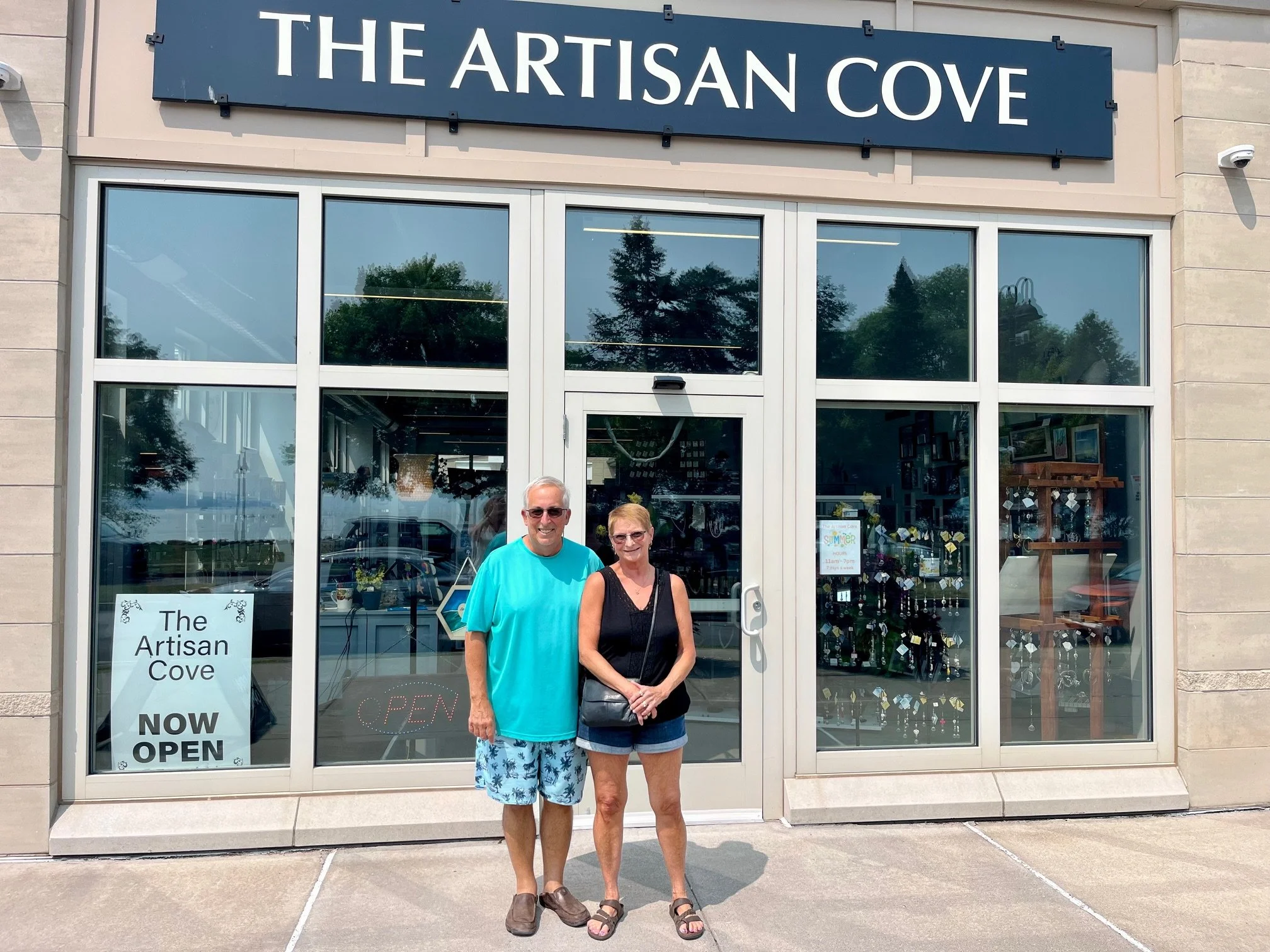 A man and woman standing outside The Artisan Cove storefront with glass windows, one sign indicating the store is now open, and a large sign above the window with the store's name.