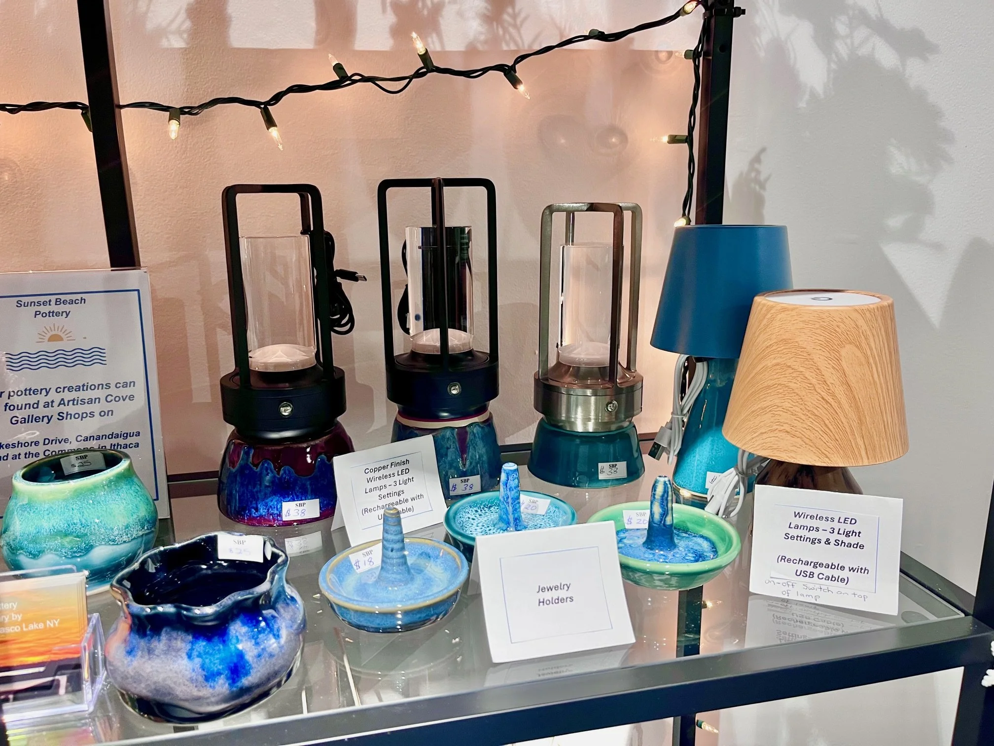 A display of ceramic pottery and lamps, including three LED lamps with wireless charging bases, decorative jewelry holders, and table lamps with shades, set on a glass table with a string of fairy lights overhead.