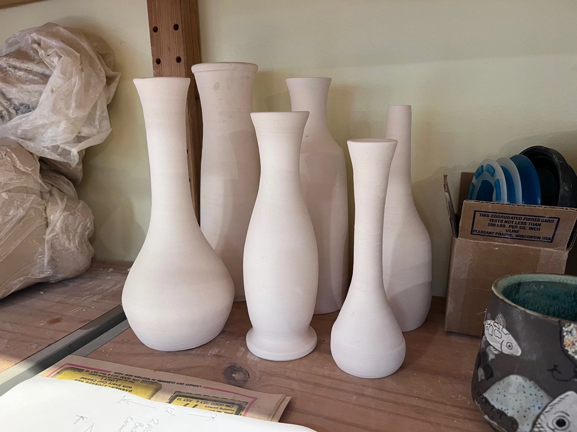 Seven unglazed ceramic vases of different shapes and sizes on a wooden workbench, surrounded by tools and supplies in a pottery studio.