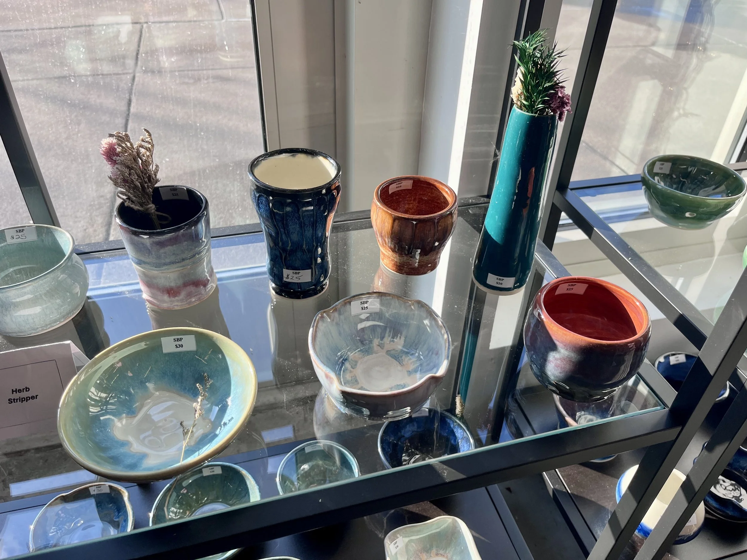 Display of colorful ceramic vases and bowls on a glass shelf near a window, with a small plant in a teal vase.