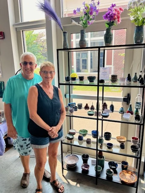 A man and woman stand next to a black metal display shelf with various colorful pottery and vases, including flowers on top, inside a bright room with large windows.