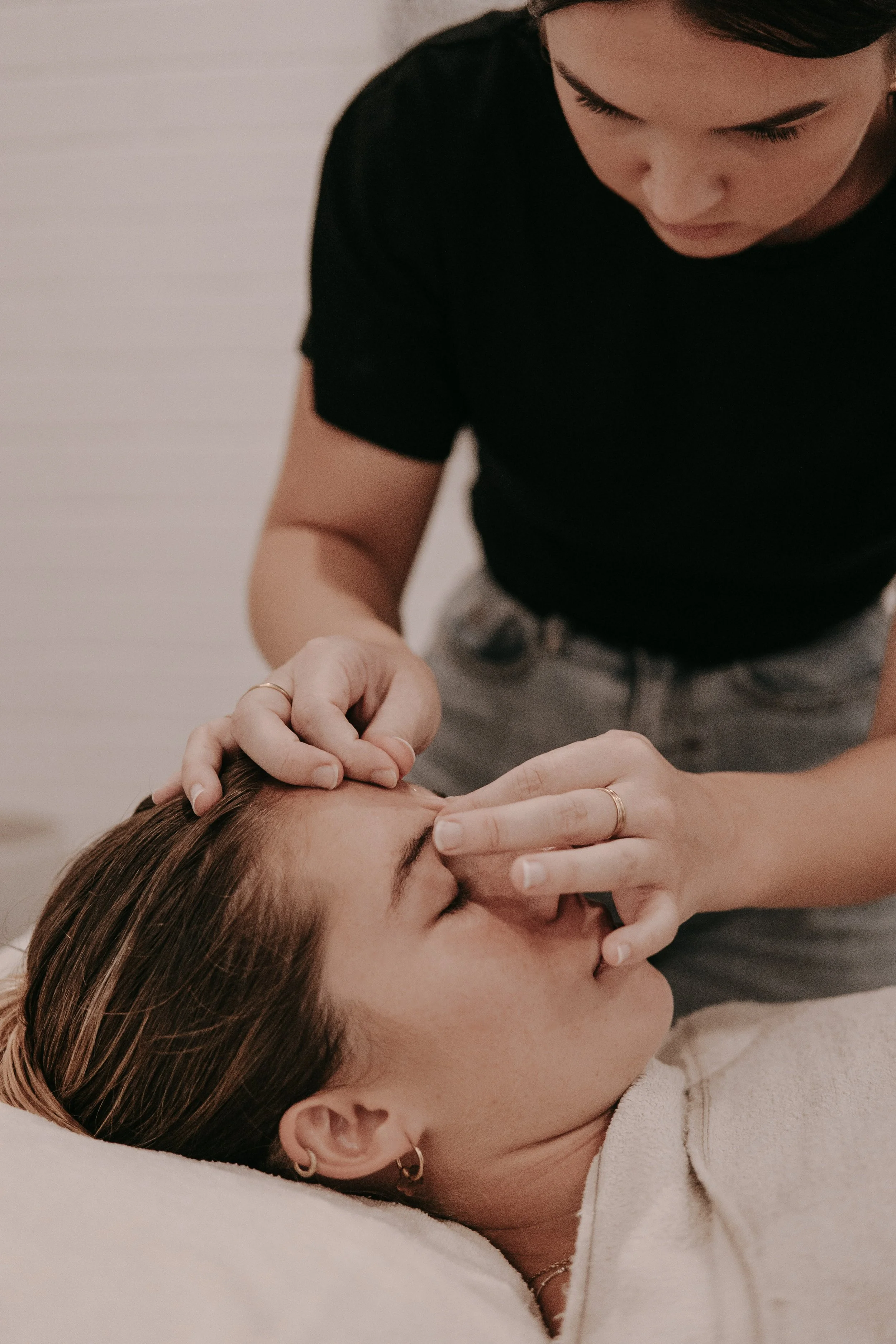 A woman receiving a facial massage or treatment while lying down with her eyes closed, a therapist standing beside her gently touching her forehead.