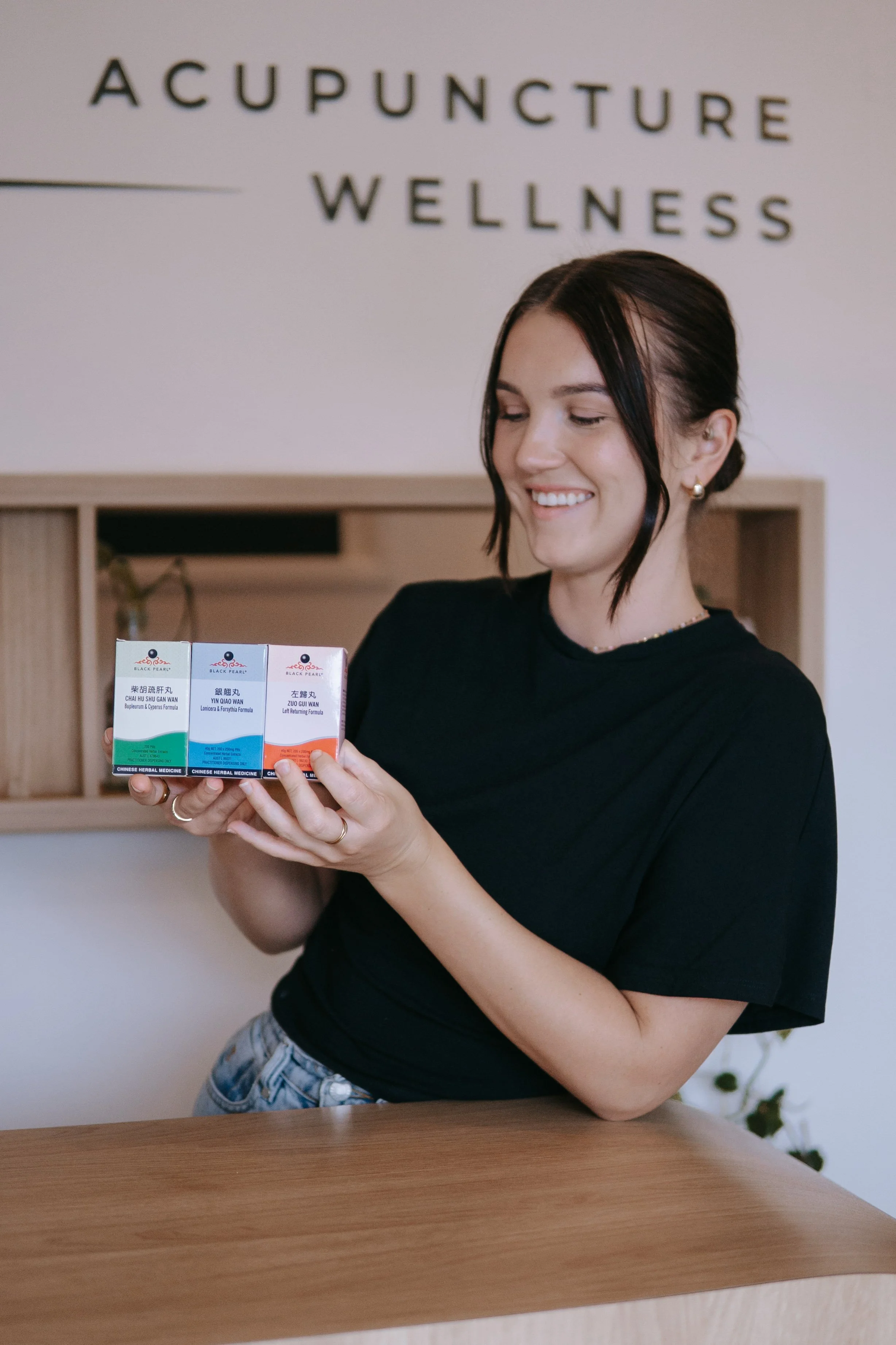 A woman with dark hair in a bun, wearing a black shirt and earrings, smiling and holding three boxes of herbal medicine, inside a wellness and acupuncture clinic.