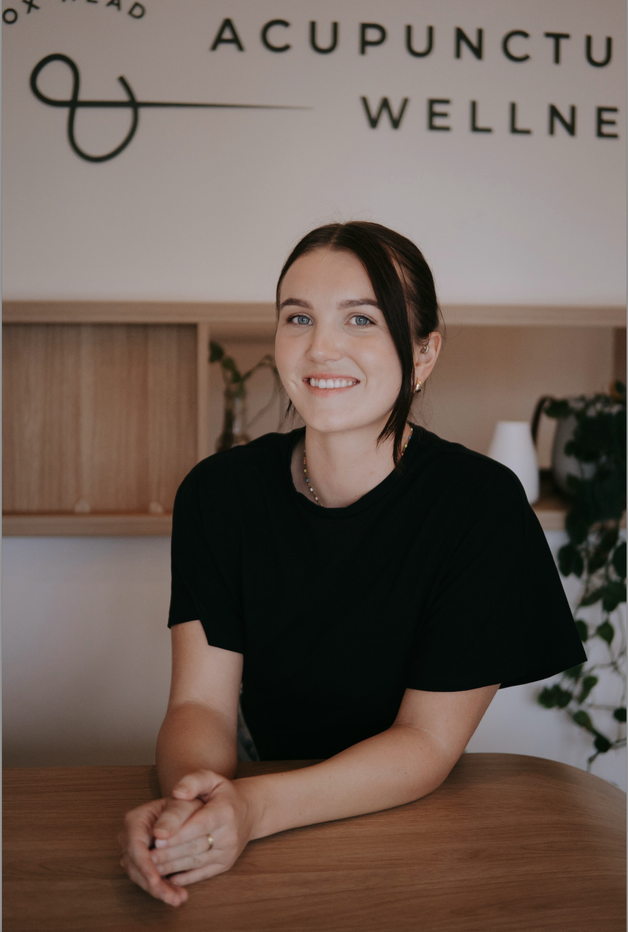 A young woman with dark hair tied back, smiling, wearing a black shirt, sitting at a wooden table in a wellness or acupuncture clinic.