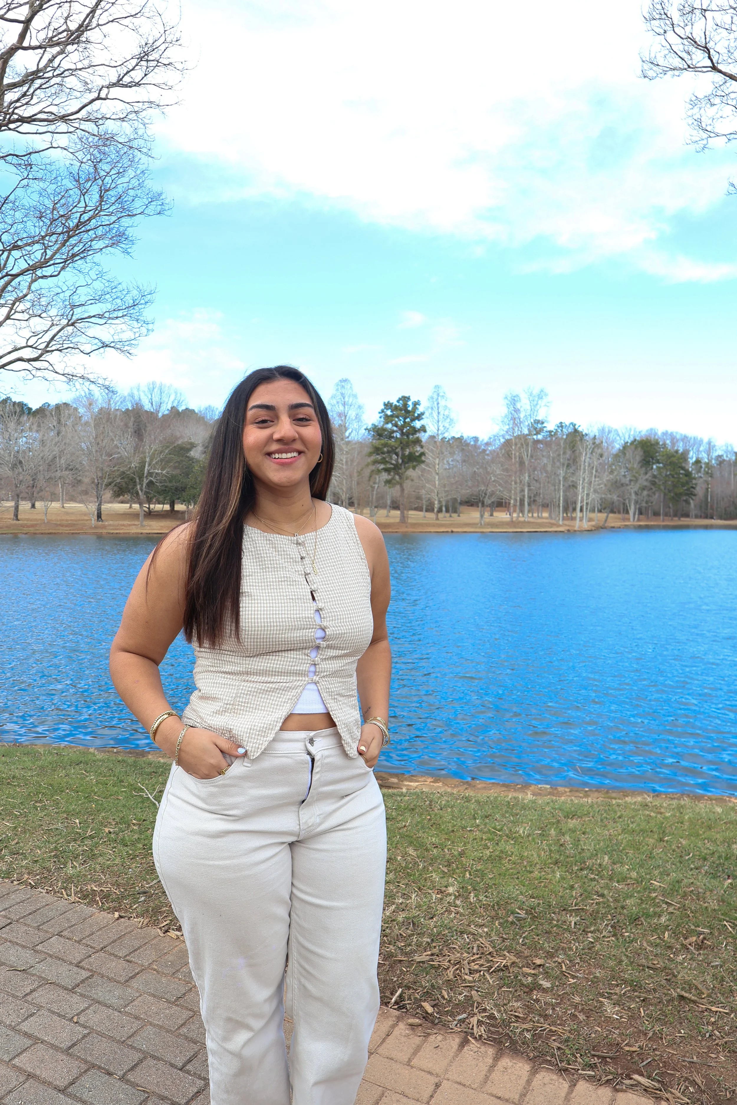 A young woman with long dark hair smiling and standing outdoors near a lake with trees in the background during daytime.