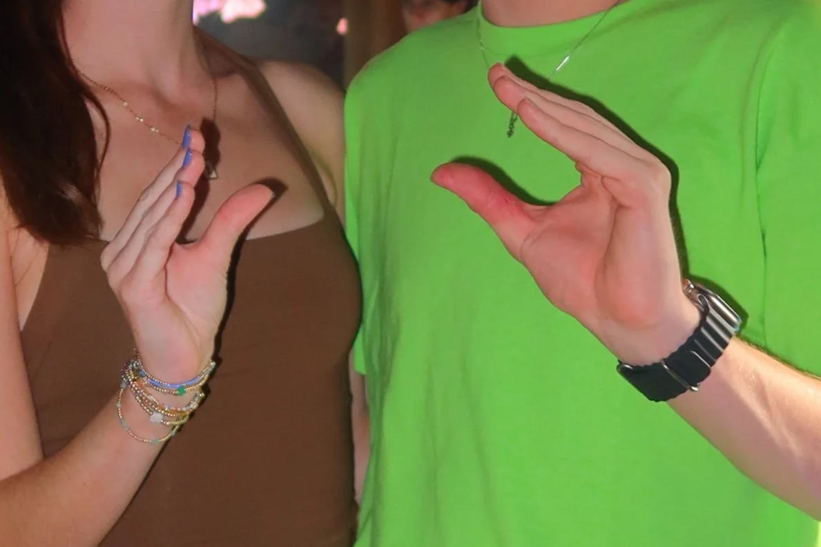 Two people with their hands raised, giving a high five or gesture. The woman on the left wears a brown sleeveless top, chains, and bracelets. The person on the right wears a bright green shirt, a watch, and a necklace.