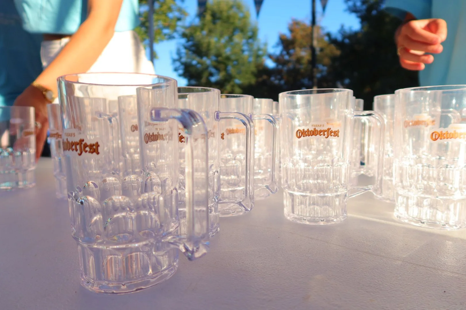 Multiple clear beer mugs with orange Oktoberfest logo on a table outdoors, with people in the background and trees against a blue sky.