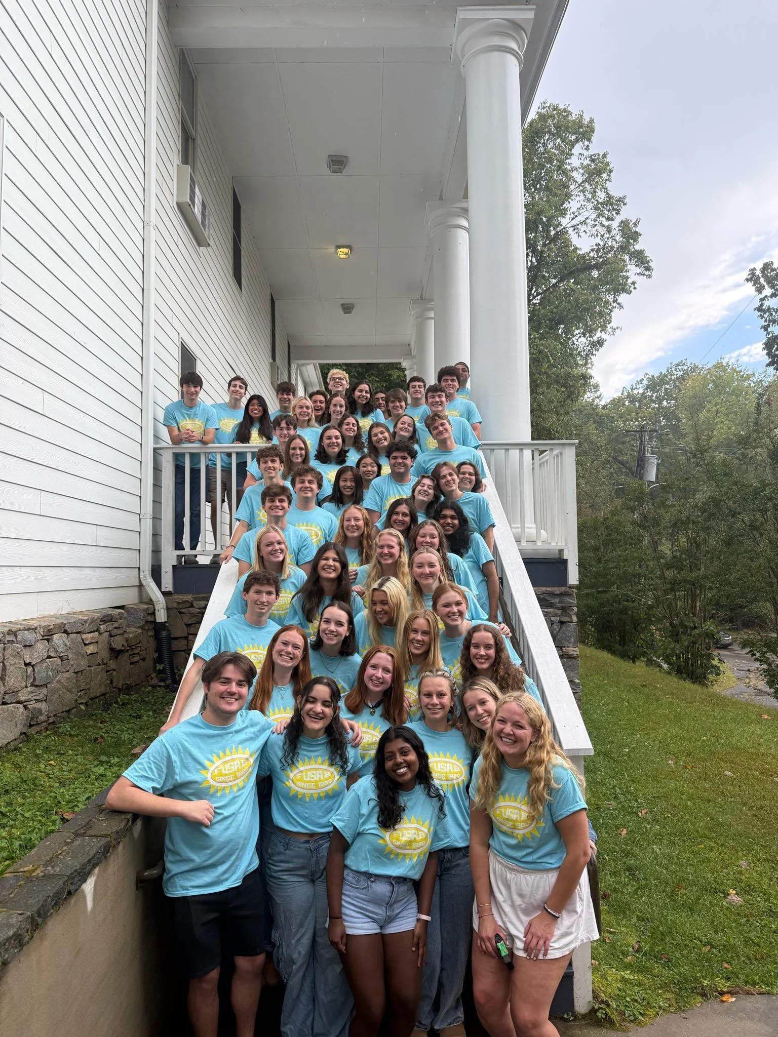 Group of young people wearing matching blue T-shirts with a yellow sun logo, standing on the steps of a white house with a porch, smiling at the camera.