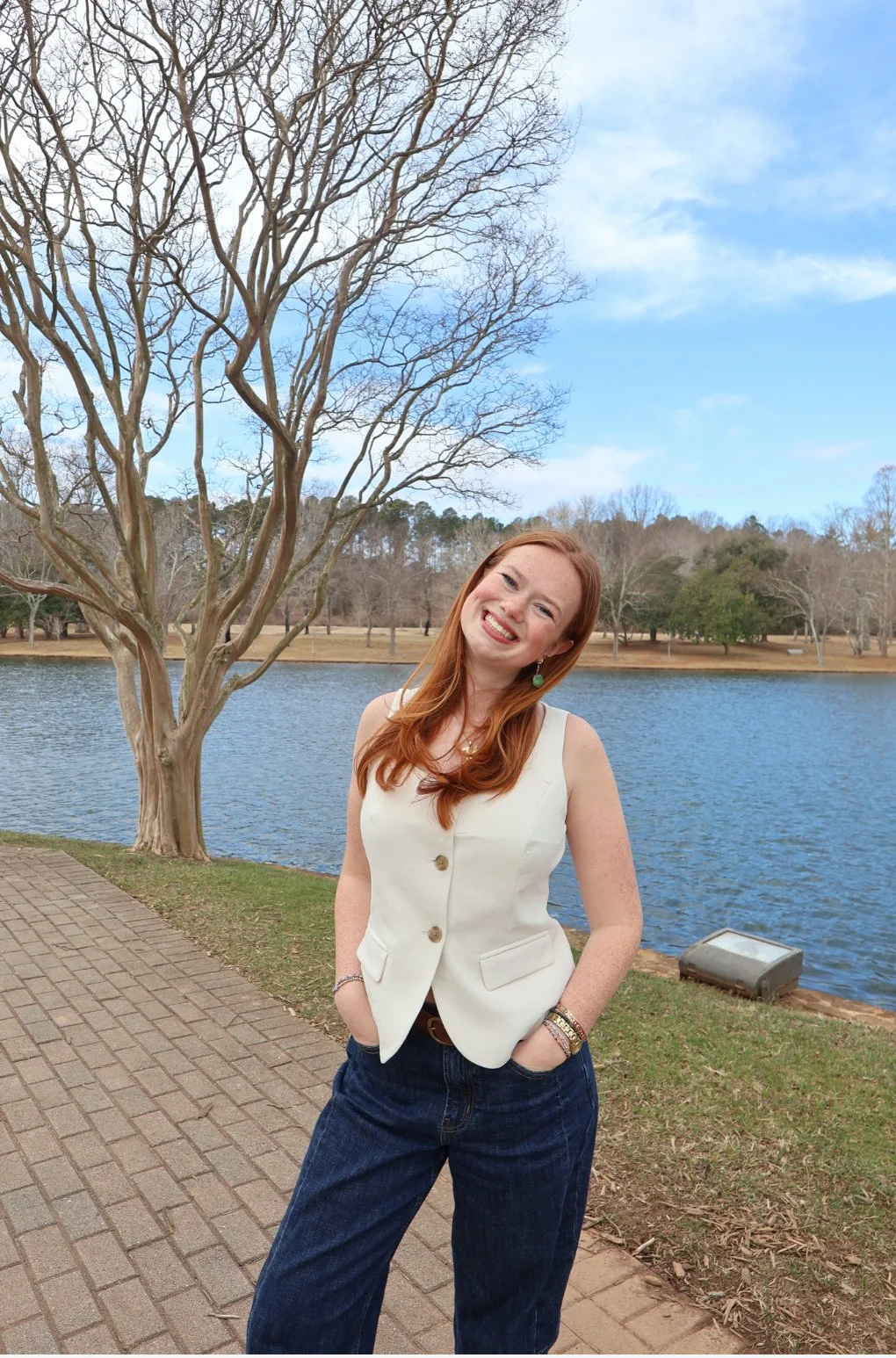 A woman with red hair smiling outdoors near a lake, wearing a sleeveless white vest and dark jeans.