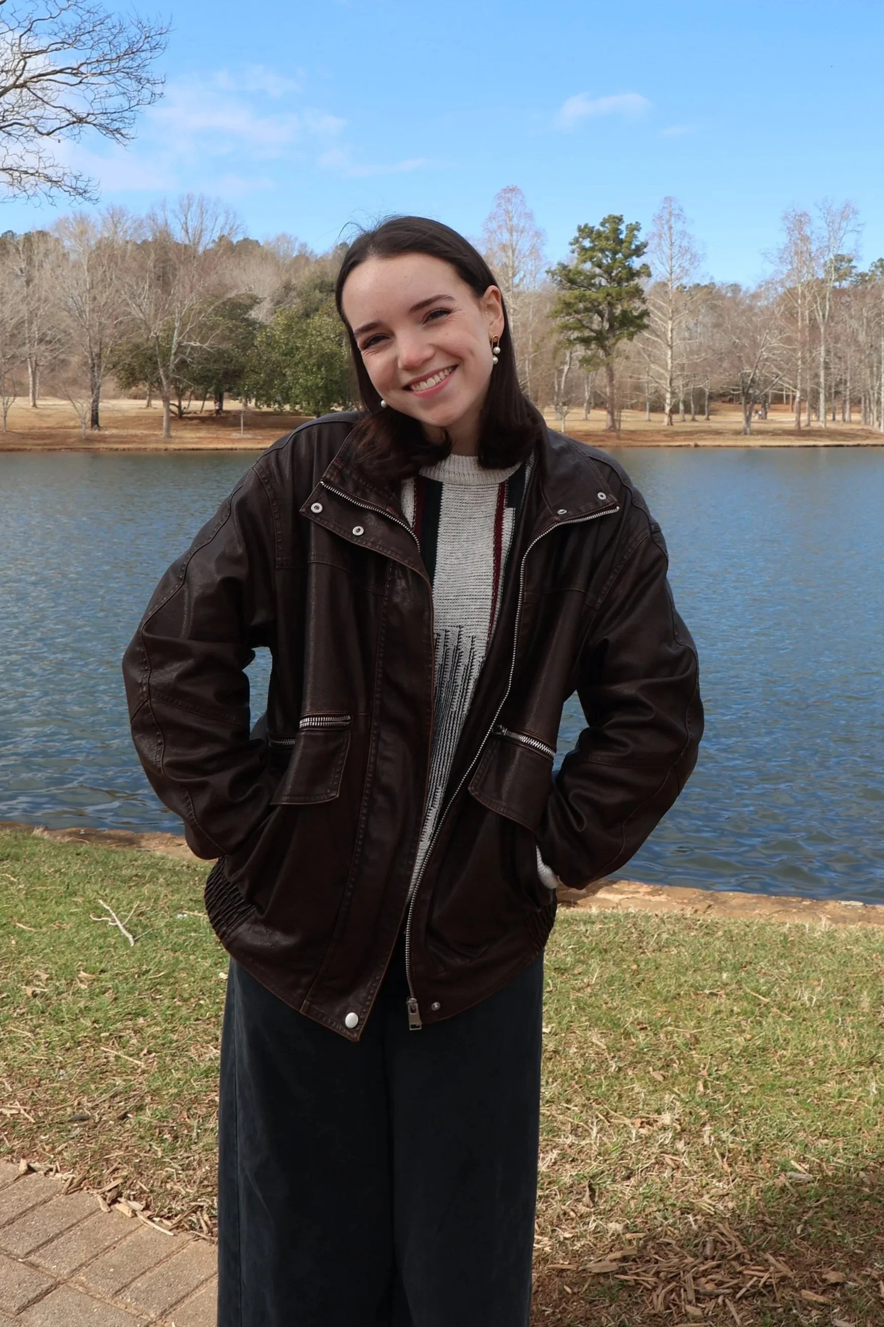 A young woman standing outdoors near a lake, smiling, wearing a dark leather jacket, sweater, and wide-leg pants, with trees and water in the background on a clear day.