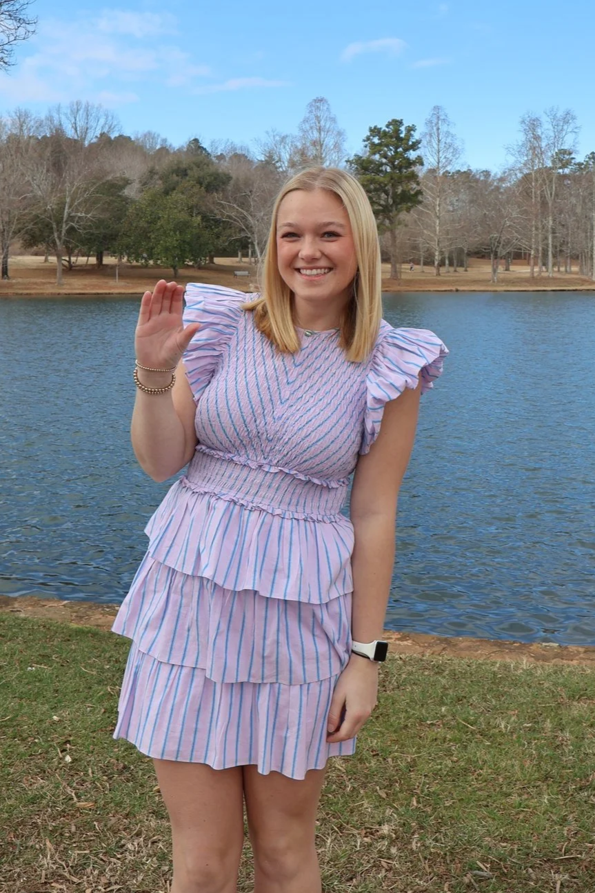A young woman with blonde hair smiling and waving near a lake, wearing a pink and blue striped dress, with trees and a partly cloudy sky in the background.