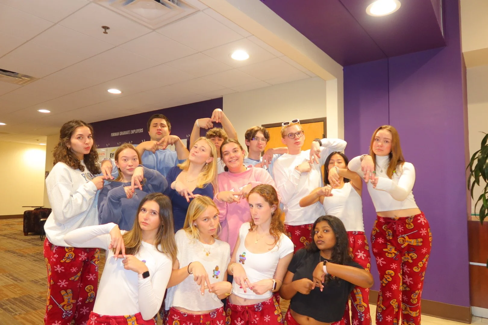 Group of young people posing indoors, wearing holiday-themed pajama pants, some with gingerbread men and snowflakes, making playful hand gestures. The background is a purple wall with a sign that reads 'Furman Graduate Employees'.