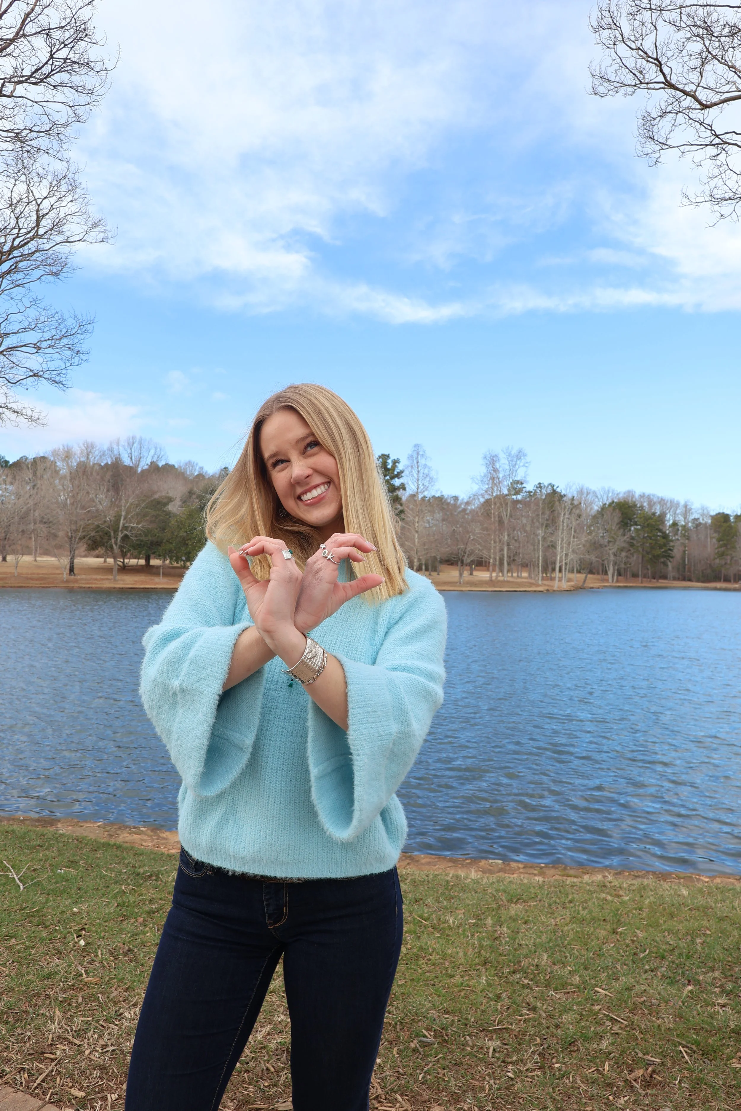 A woman with blonde hair smiling and making a heart gesture with her hands standing outdoors near a lake with a blue sky and some trees in the background.