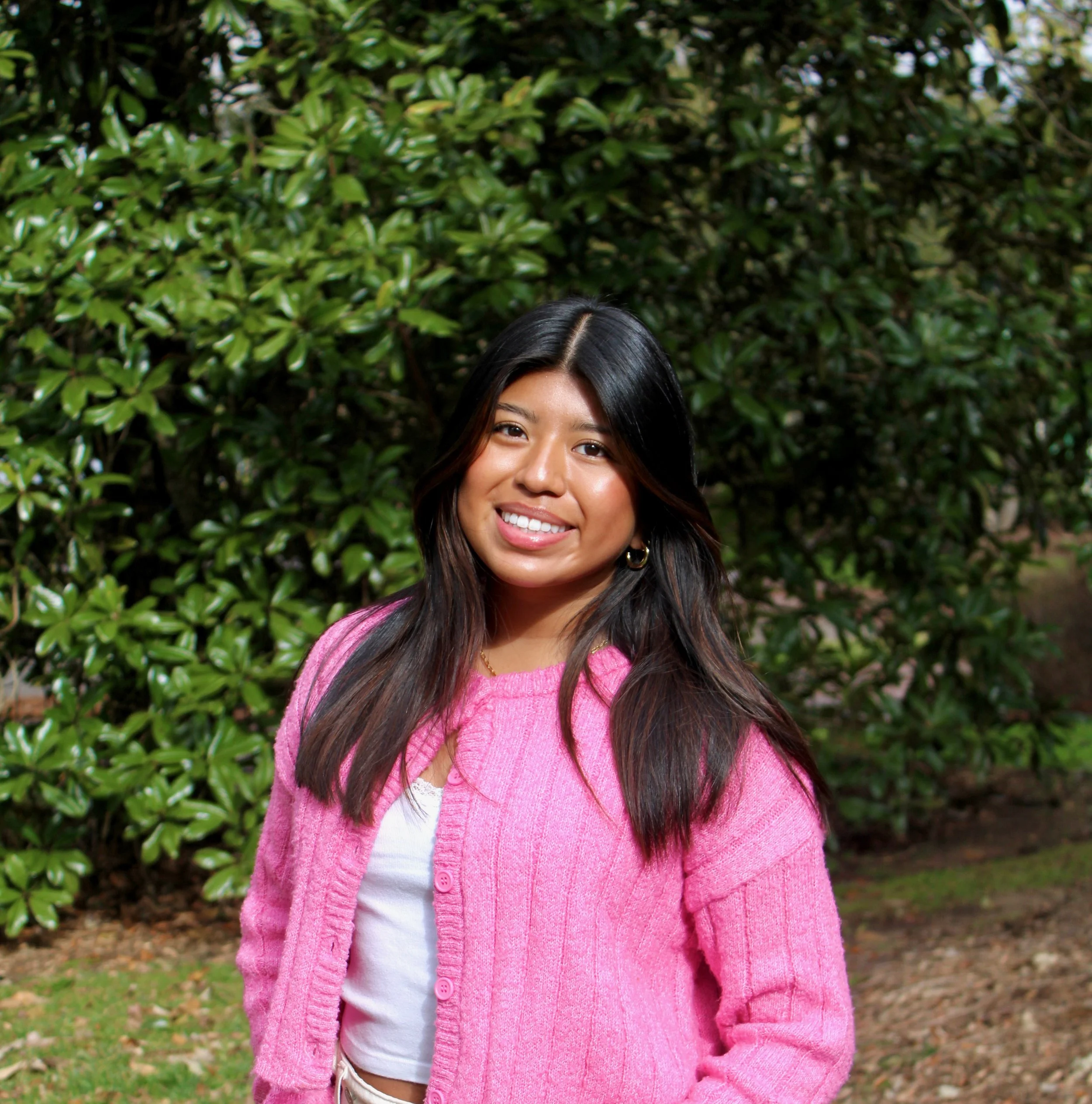 A woman with long dark hair, wearing a pink cardigan over a white top, smiling outdoors with green bushes in the background.