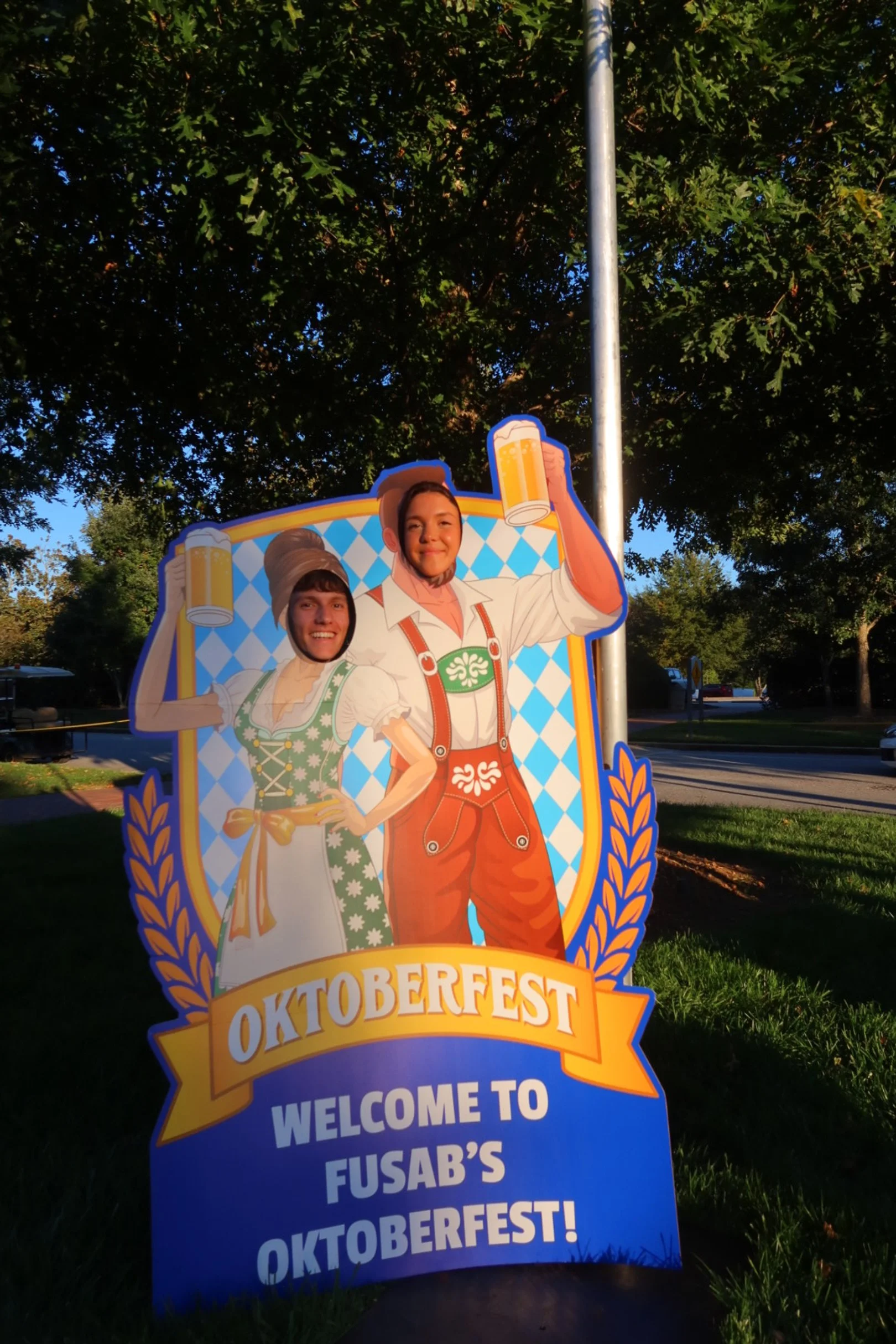 Decorative sign for Oktoberfest featuring cartoon characters in traditional German clothing, holding beer mugs, with text welcoming visitors to Fusab's Oktoberfest, set outdoors near trees and a street.