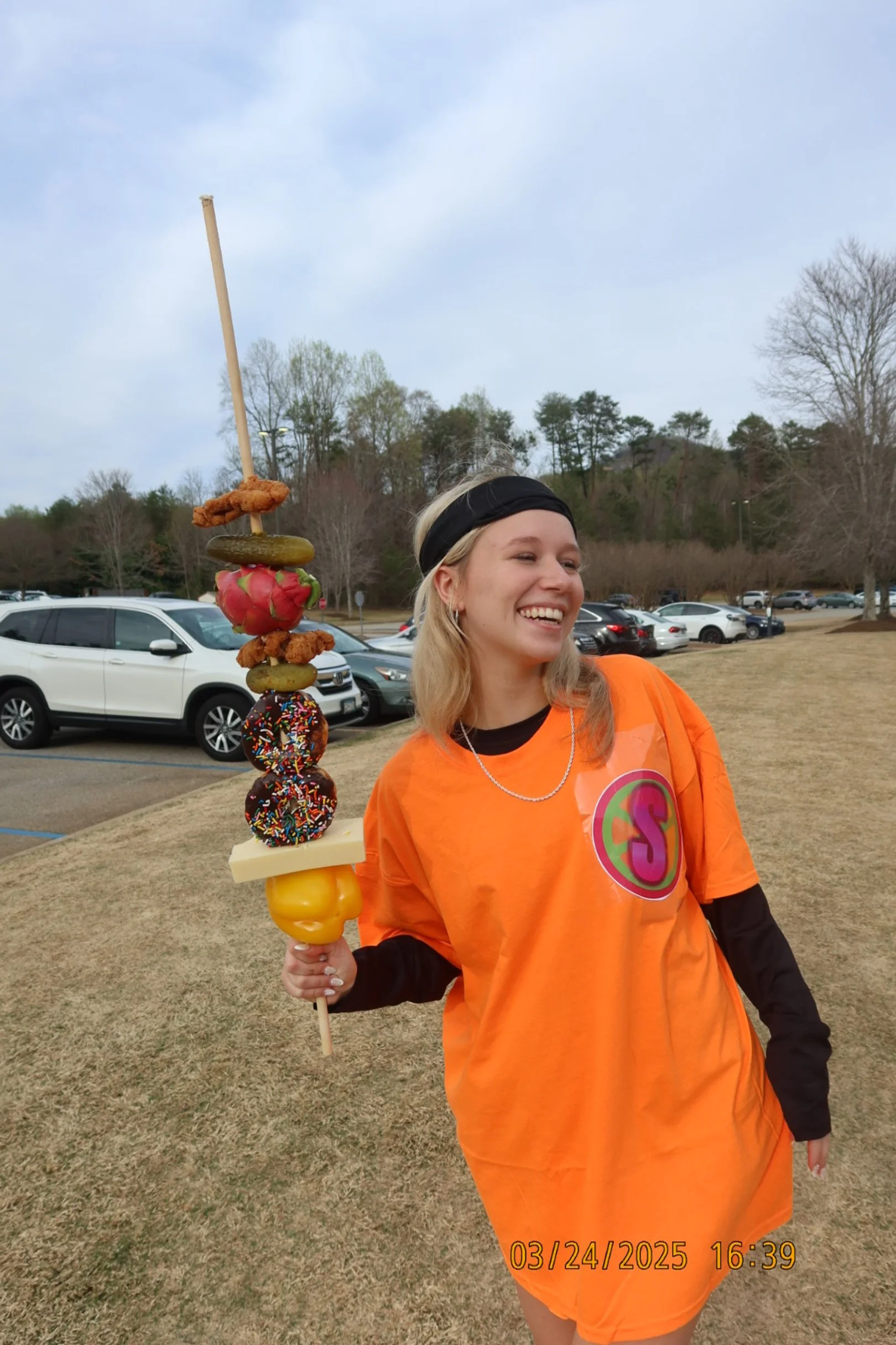 Young woman smiling and holding a skewer with various snacks including fried chicken, pickles, a strawberry, sprinkled donuts, and yellow bell pepper, standing outdoors in a park with cars and trees in the background.