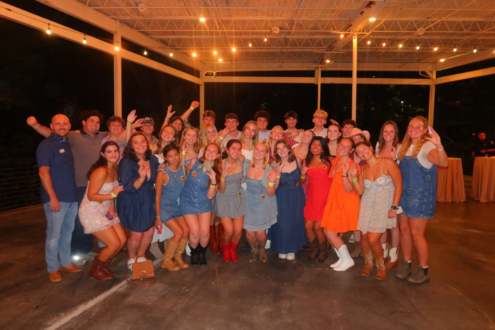 Group of young adults at a social event, wearing casual and colorful outfits, posing indoors with smiles and various hand gestures.