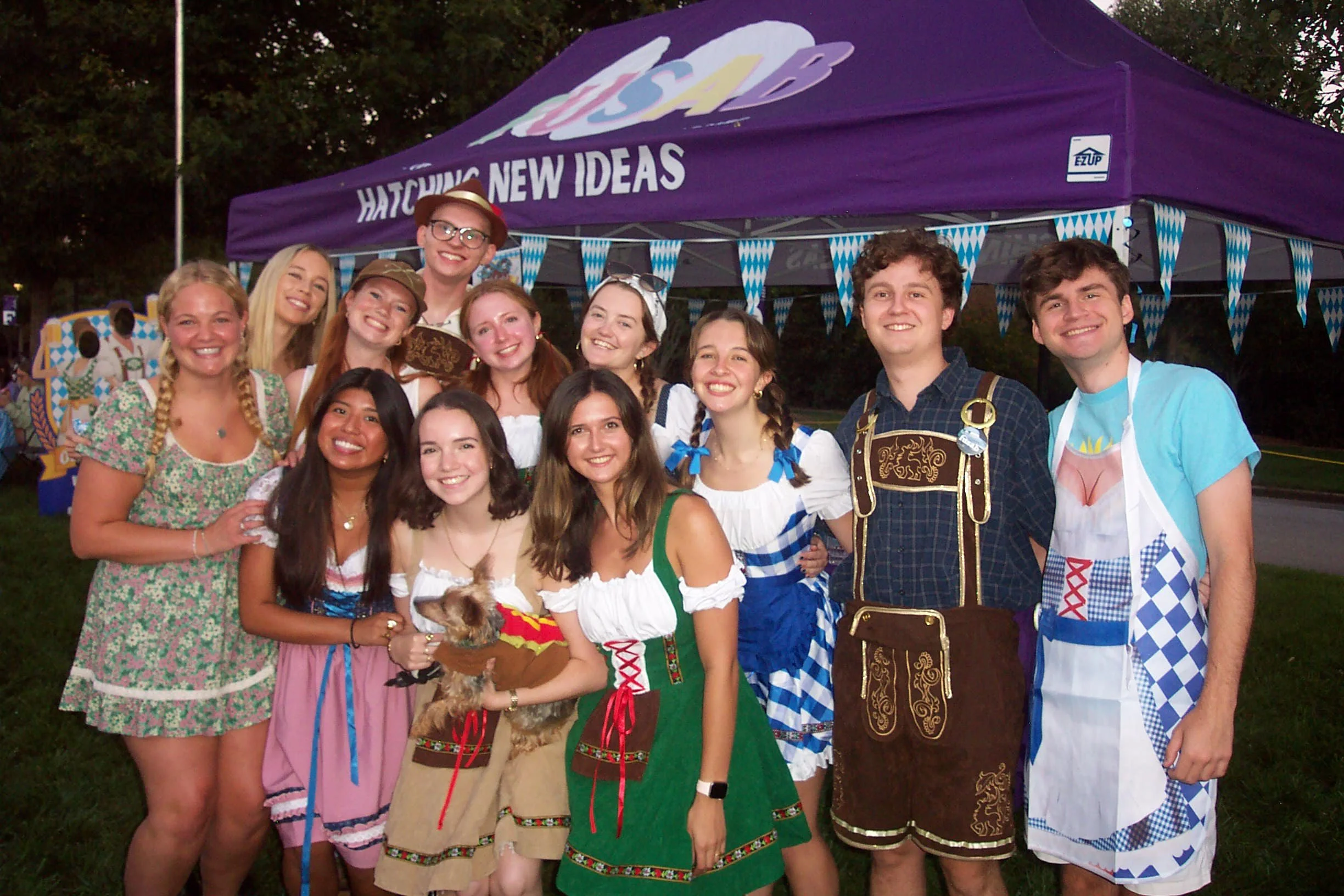 Group of young people celebrating at an outdoor festival, many dressed in traditional Bavarian or Oktoberfest costumes, standing in front of a purple tent with banners.