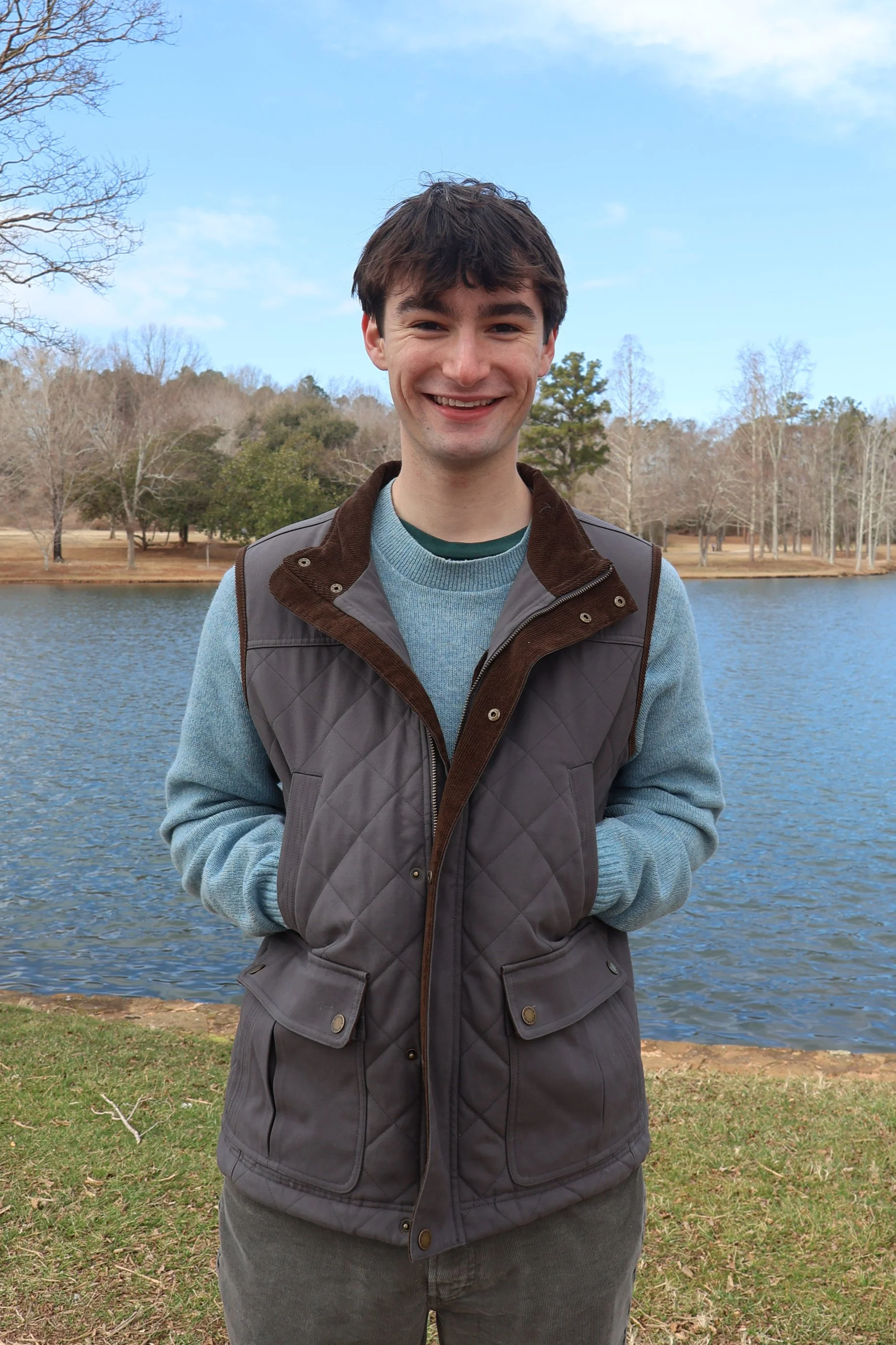 Young man smiling outdoors near a lake with trees in the background.