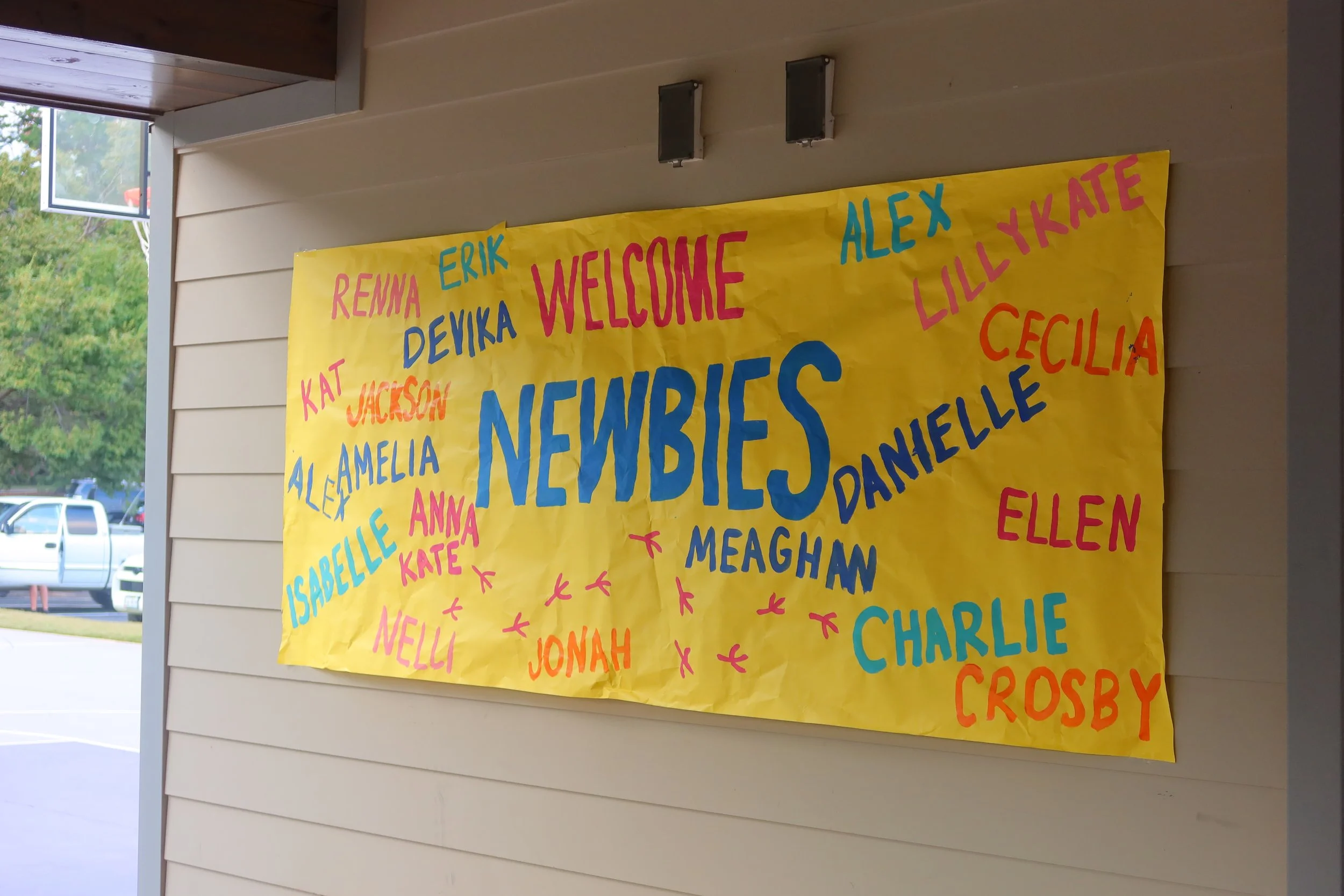 A decorated welcoming poster on a wall with the word 'NEWBIES' in large blue letters and surrounded by names of children in different colors, including Renna, Erik, Alex, Katie, Cecilia, Ellen, Charlie, Crosby, Meaghan, Jonah, Nelli, Anna, Kate, Isab