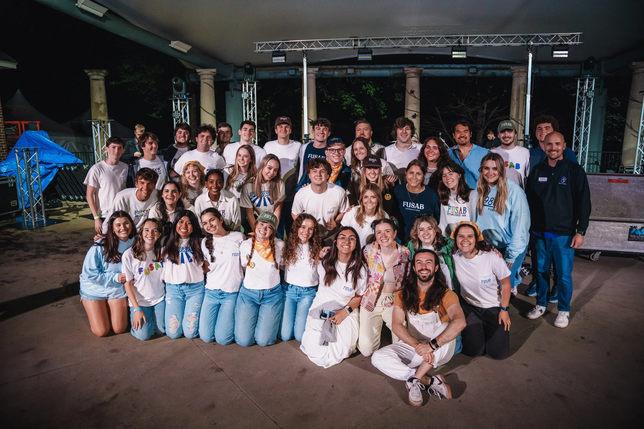 Group of smiling young people and adults posing for a group photo outdoors at night on a stage with lighting and columns in the background.