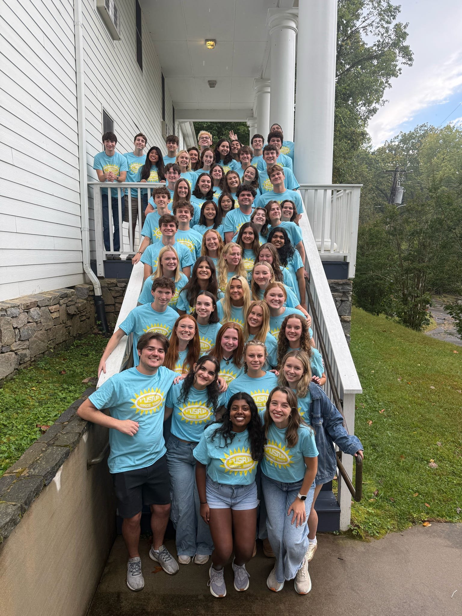 Group of young people standing on an outdoor staircase, wearing matching light blue T-shirts with a sun design and the word 'FUSAB' printed on them, smiling and posing for a photo.