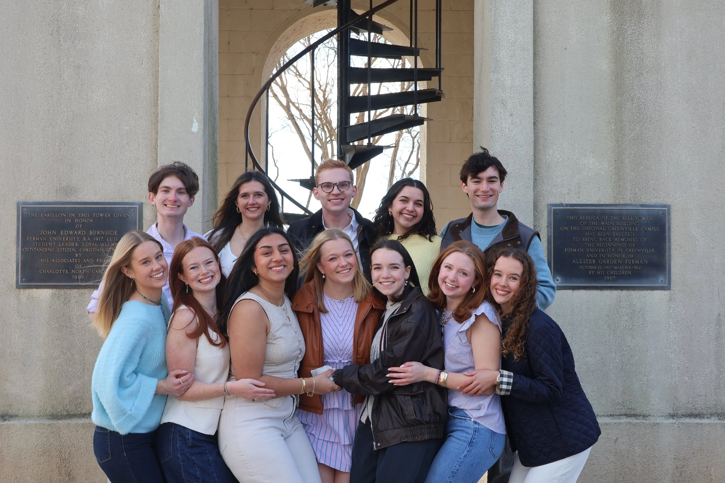 Group of young adults smiling and posing for a photo outside in front of an architectural wall with plaques, a spiral staircase, and trees in the background.