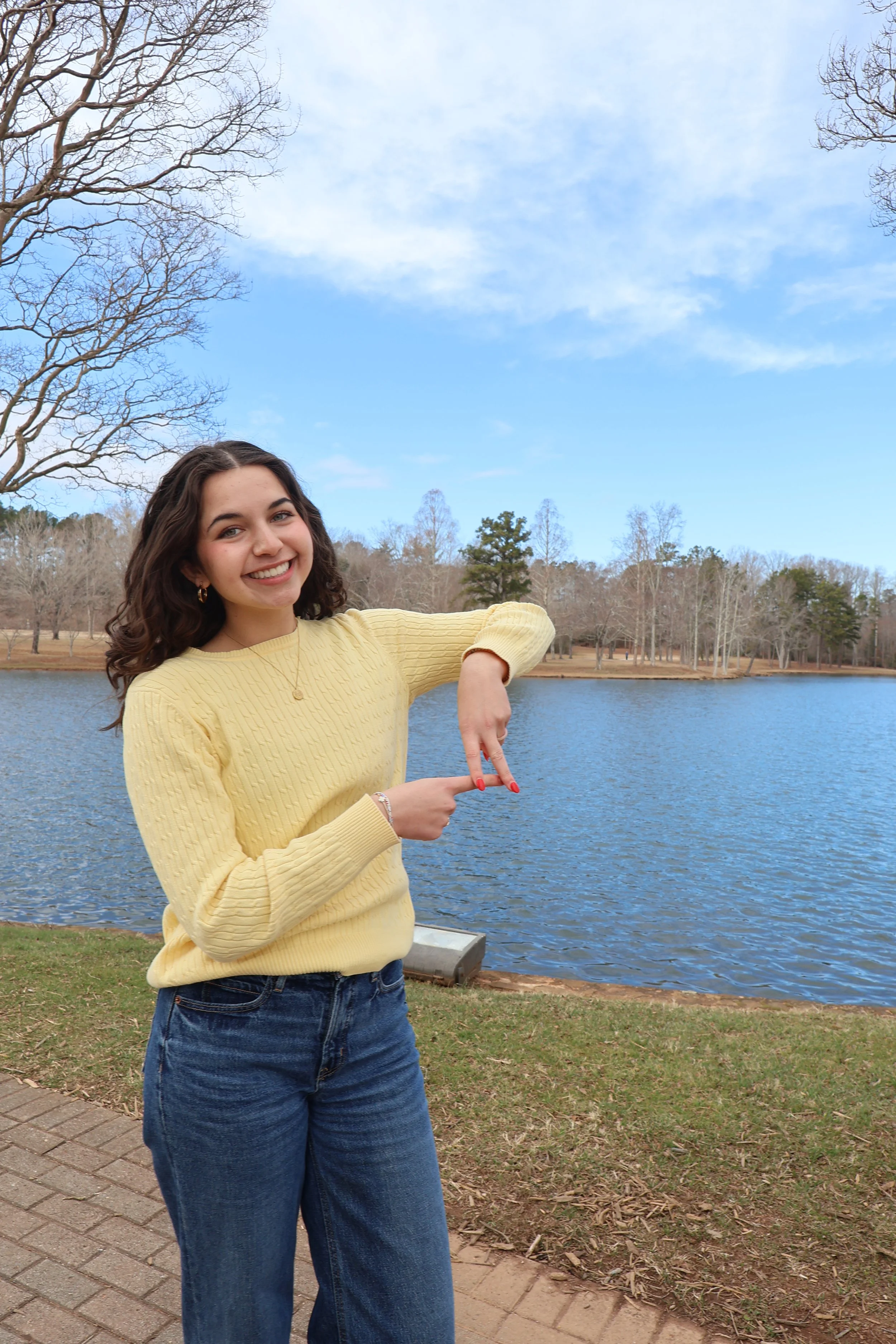 A young woman in a yellow sweater and blue jeans smiling and pointing at a lake with a natural landscape of trees in the background.