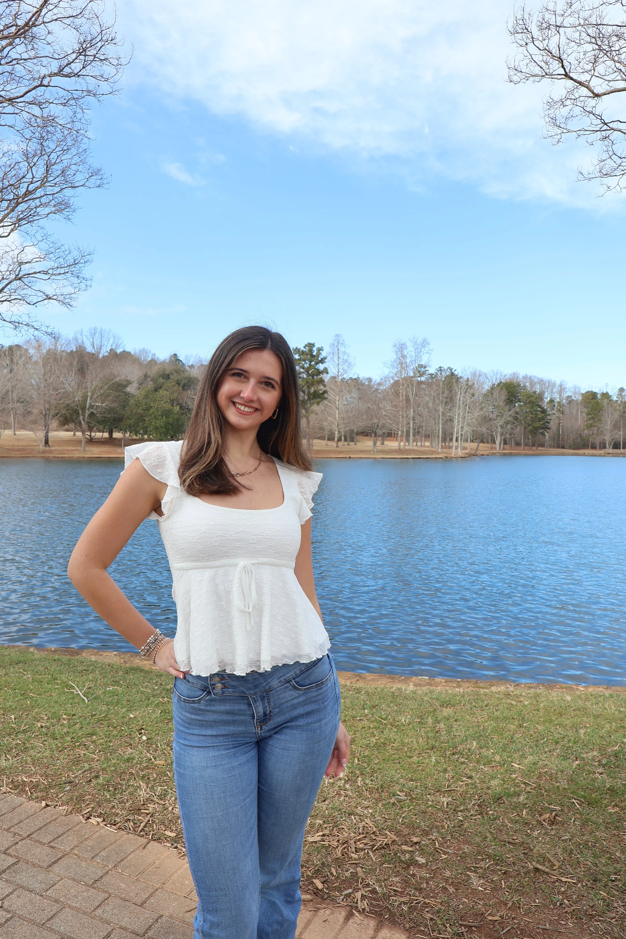 A young woman with long brown hair wearing a white top and blue jeans standing outdoors near a lake with trees in the background on a partly cloudy day.