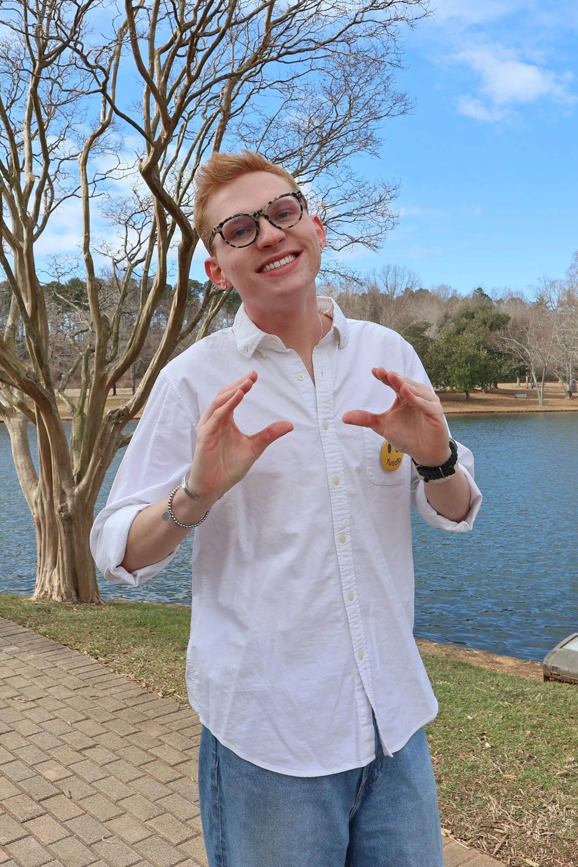 A young man with glasses and a white shirt standing near a lake with bare trees in the background, smiling and posing outdoors on a cloudy day.