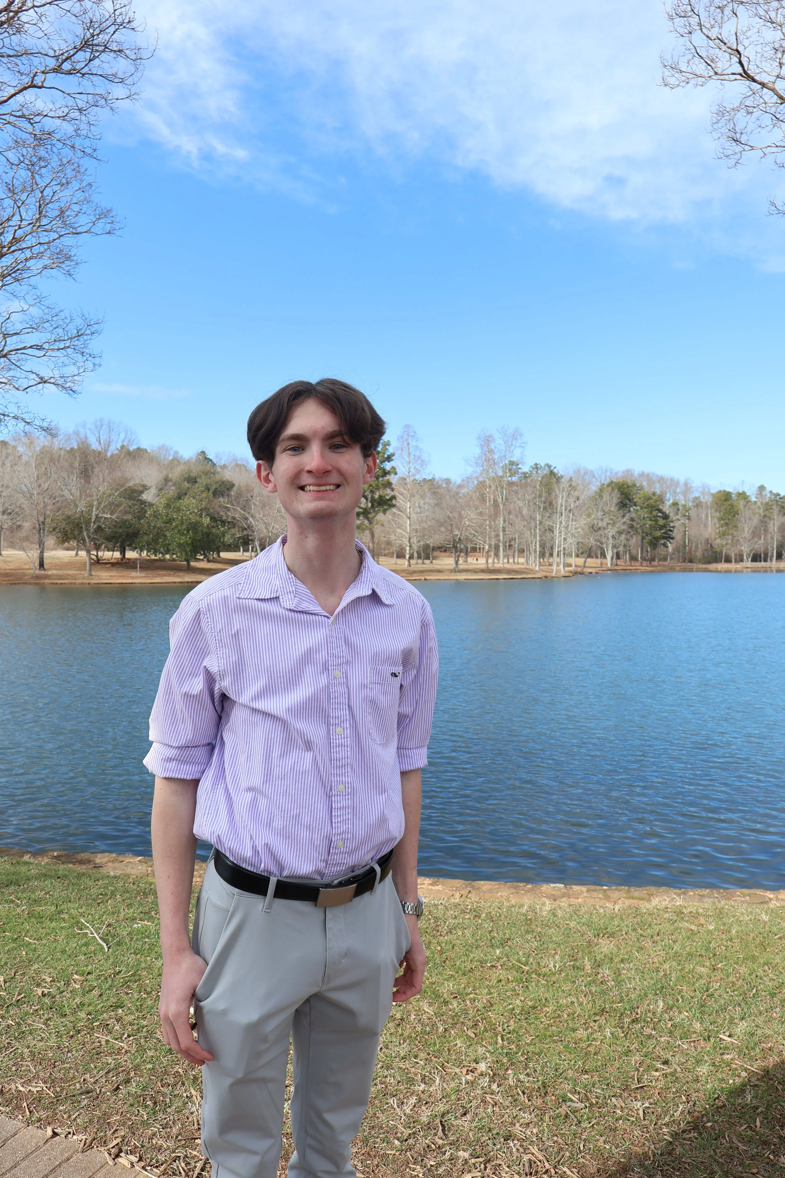 A young man in a purple striped shirt and beige pants standing by a lake with trees in the background, under a partly cloudy sky.