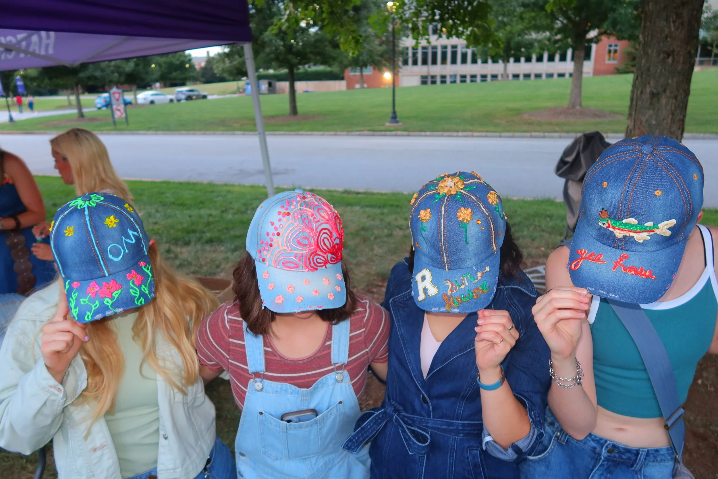 Four women with decorated denim caps covering their faces, standing outdoors under a tent.