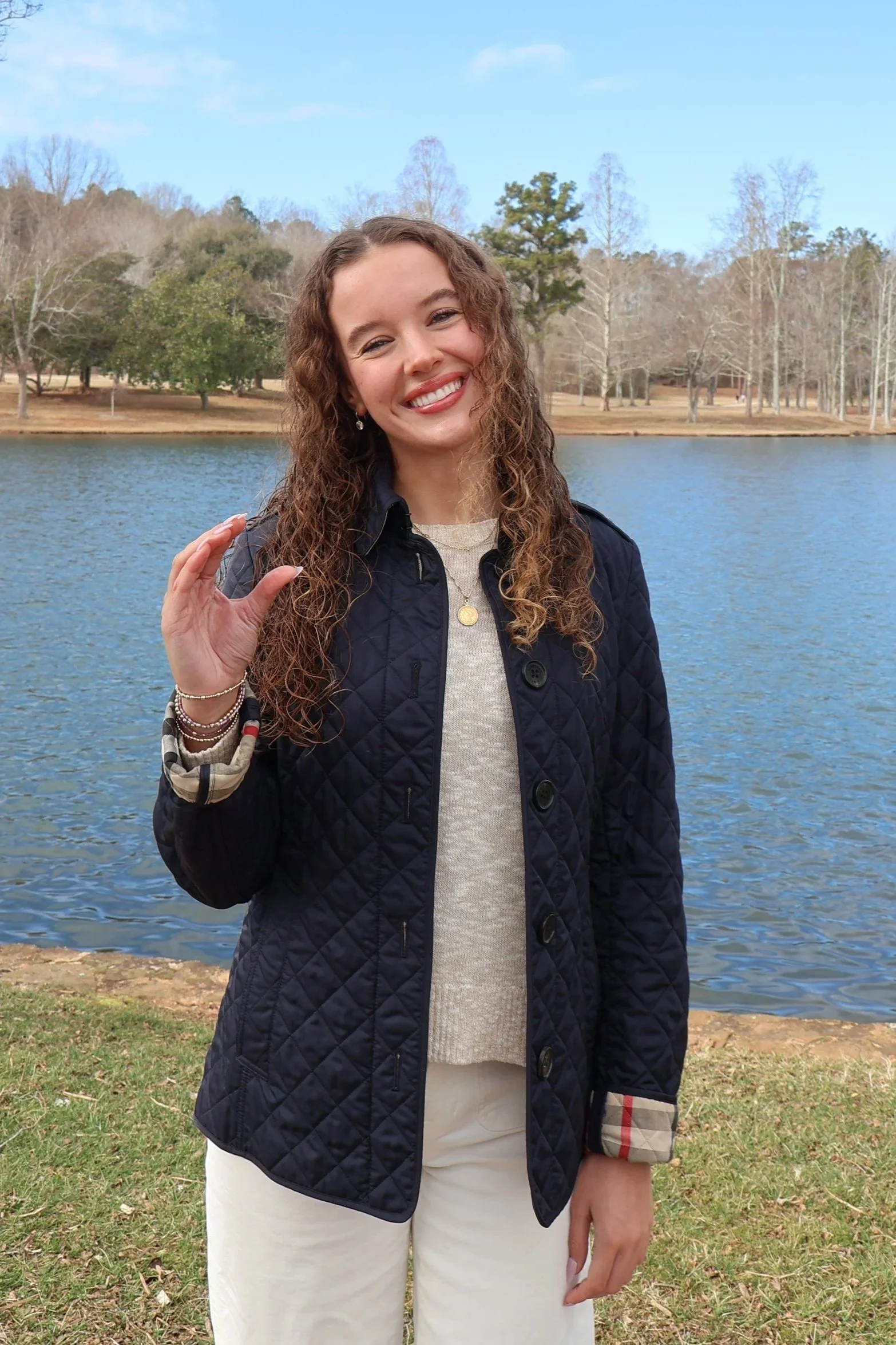 A young woman with curly brown hair, smiling, standing near a lake during daytime, wearing a navy quilted jacket, beige sweater, and white pants, with trees and a clear blue sky in the background.