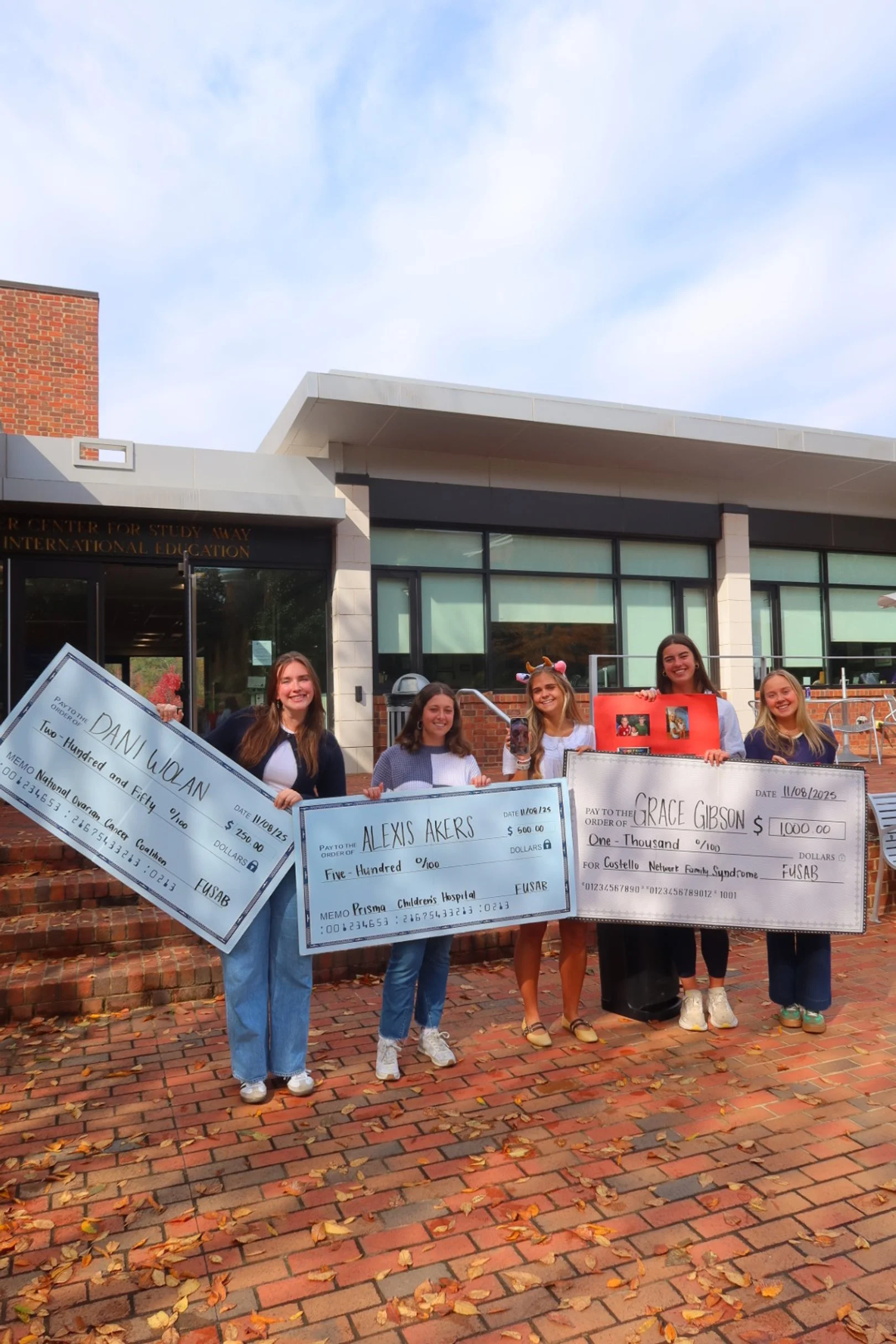 Five young women standing outside a building, holding oversized checks for different amounts made out to various recipients. They are smiling and dressed casually, with some wearing animal horn headbands. The ground is covered with fallen autumn leav