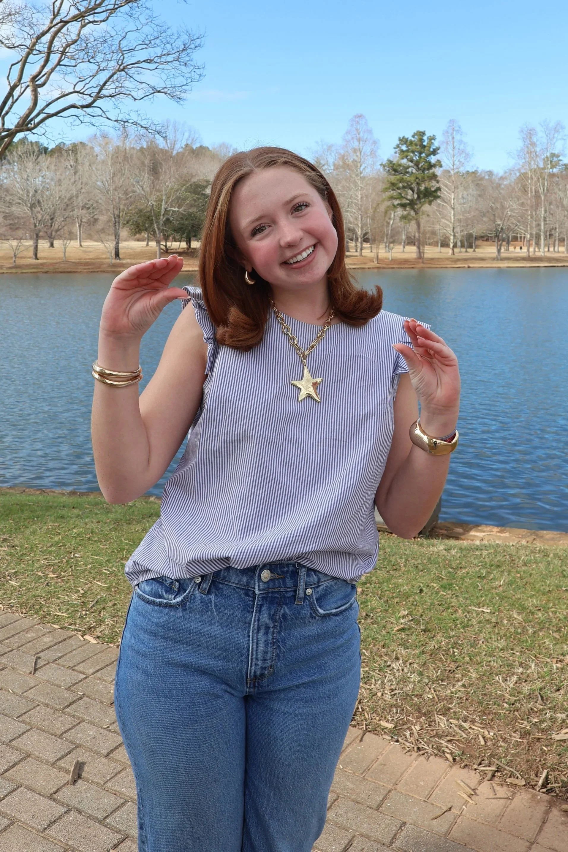 A young woman with red hair smiling outdoors by a lake, wearing a light blue striped sleeveless top, blue jeans, gold jewelry, and a star necklace, with trees and a blue sky in the background.