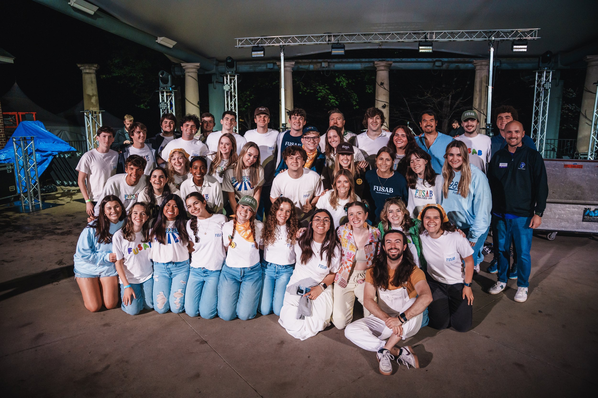 Group of young people and adults posing for a photo at an event stage with lighting equipment and metal trusses, some wearing white and blue shirts with a conference or organization logo, inside a covered venue.