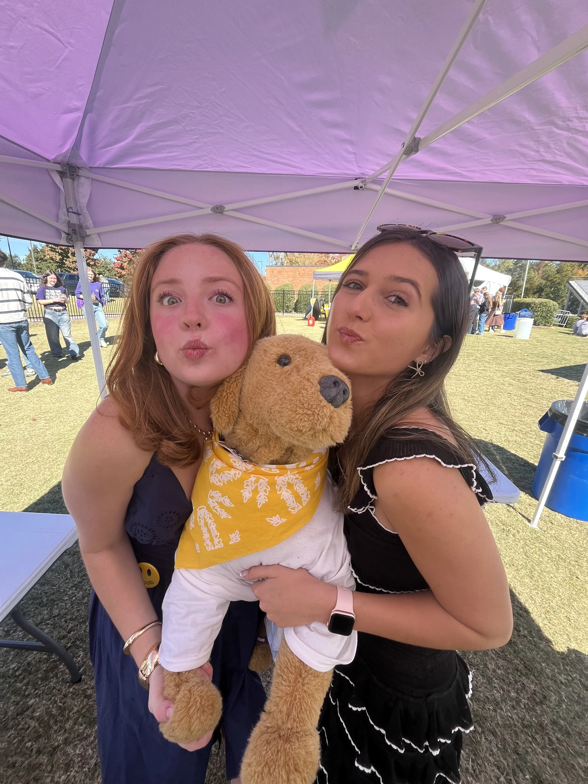 Two FUSABers with a stuffed dog, Rowdy, posing for a photo under a purple tent.
