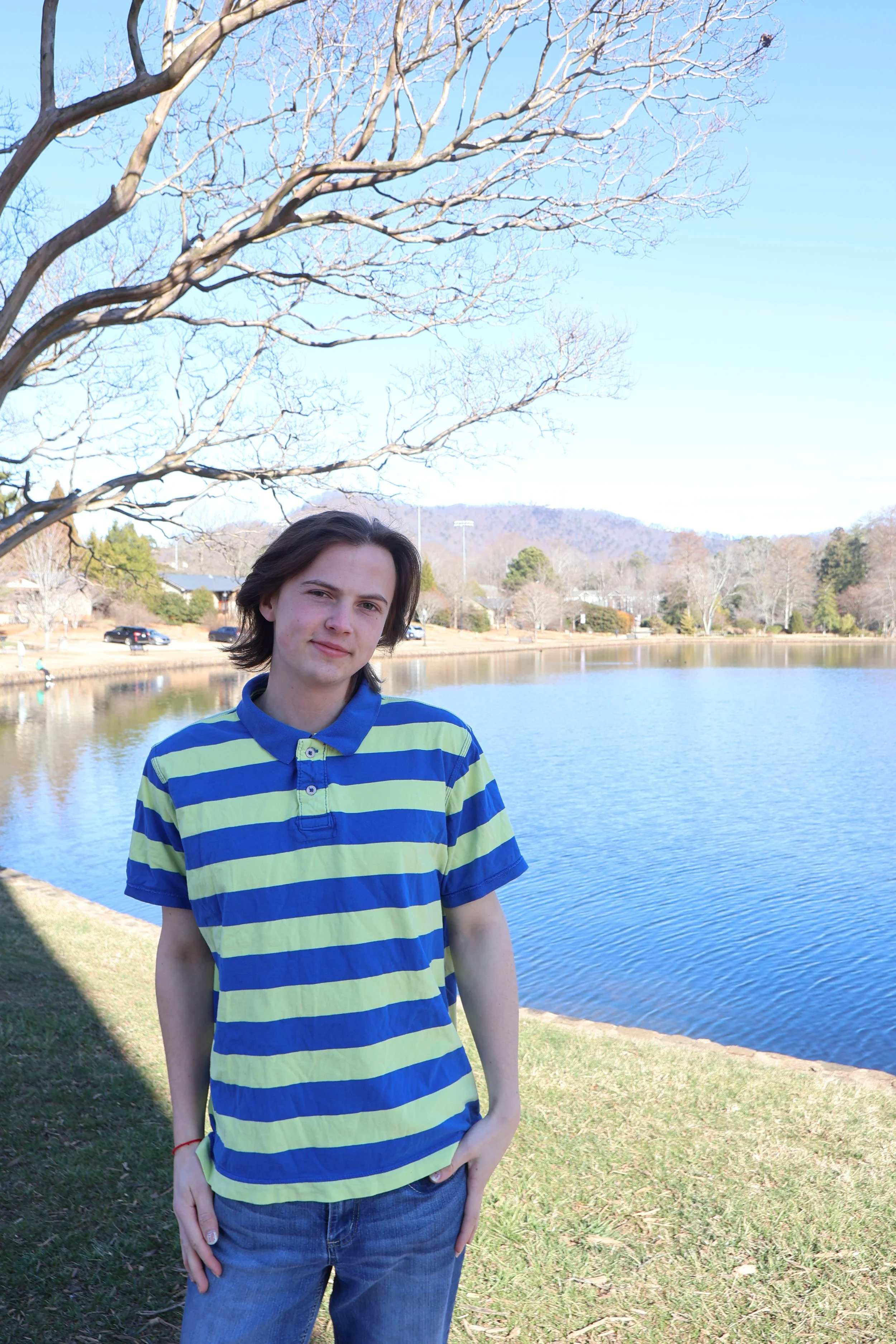 A young man wearing a yellow and blue striped polo shirt and jeans standing near a lake with some trees and mountains in the background on a clear, sunny day.