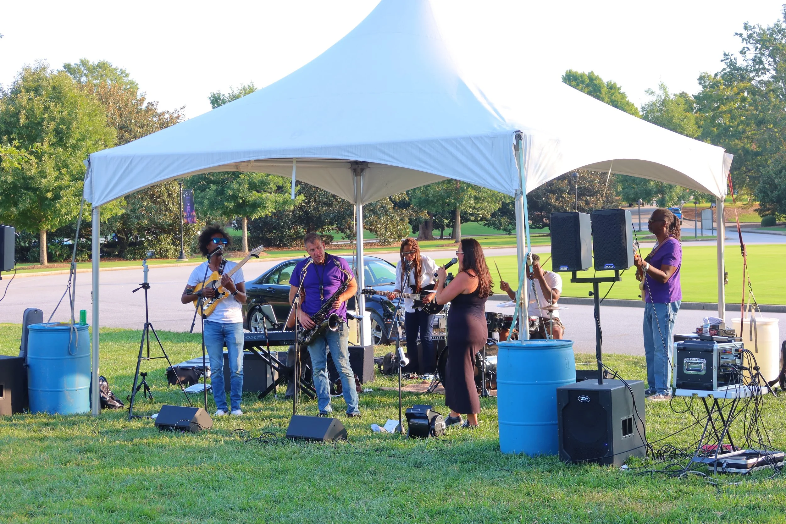 A band of six musicians performing under a white canopy tent on a grassy area in a park, with trees and a street in the background. The band includes guitarists, saxophonist, drummer, and singer, with music equipment and speakers around them.