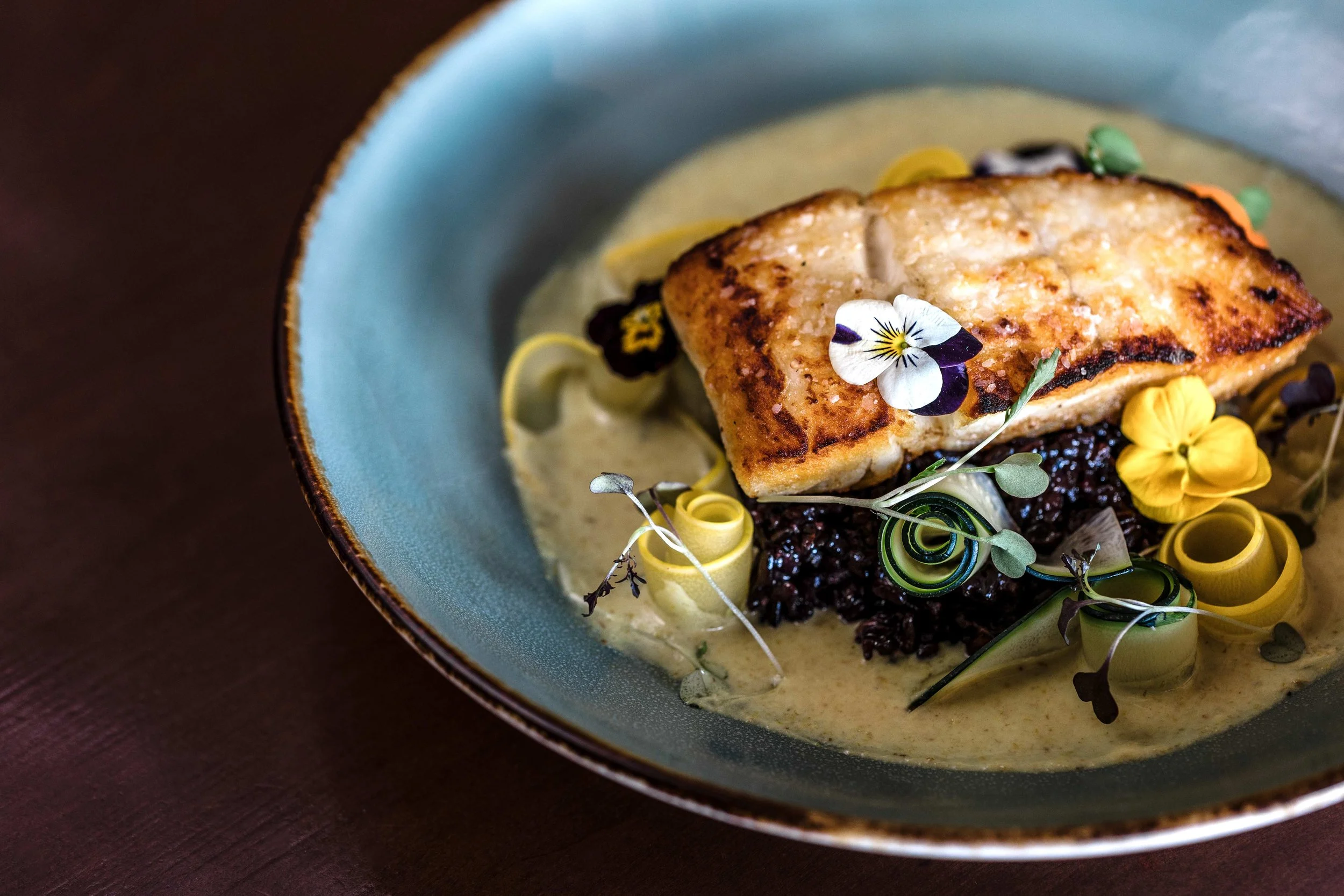 A plated dish featuring a cooked fish fillet garnished with edible flowers and herbs, served on a bed of yellow and dark-colored vegetable ribbons and dressed with a creamy sauce in a light blue bowl with a dark brown background.