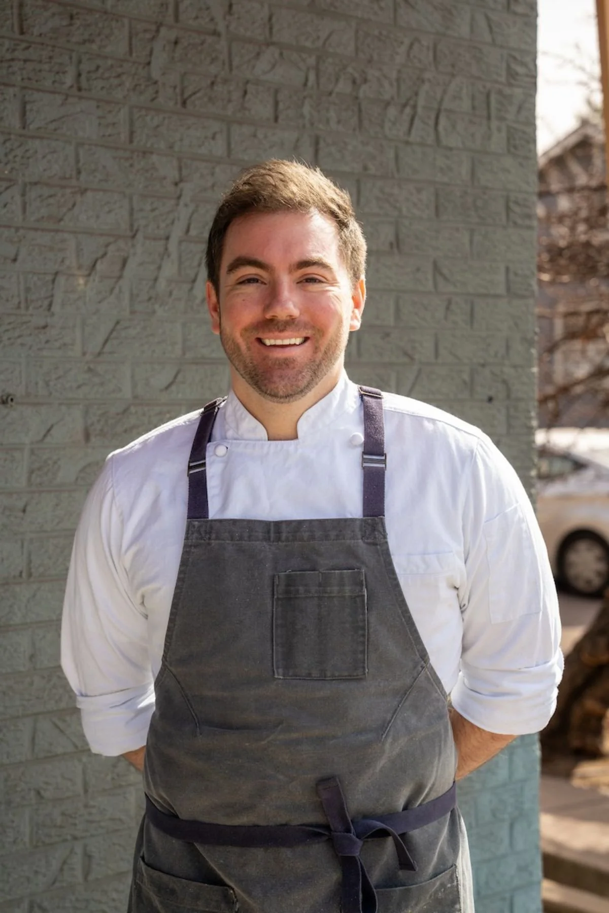 Confident male chef smiling outdoors, wearing a white chef coat and dark apron, standing in front of a brick wall.