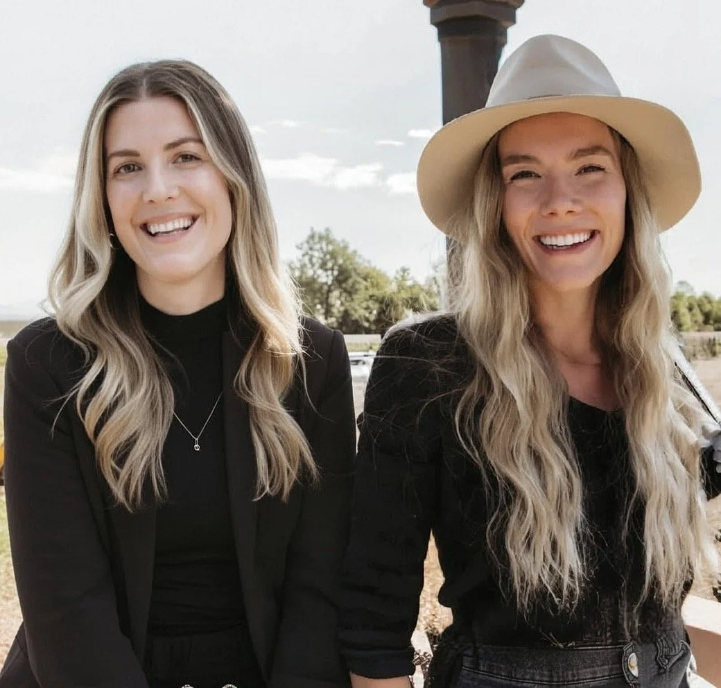 Two women smiling outdoors, one wearing a black blazer and the other wearing a white hat, with a background of trees and a cloudy sky.