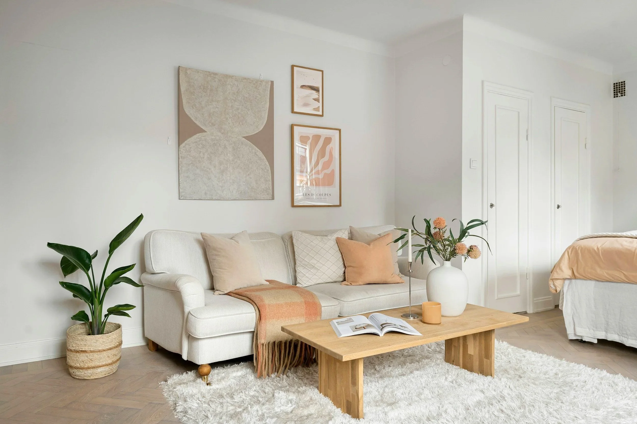 A living room with a white upholstered sofa with cream and beige cushions, a wooden coffee table with a large white vase holding pink flowers, a candle, and an open book, a green potted plant in a woven basket, framed abstract art on the wall, a fluffy white rug, and a bed partially visible in the background.